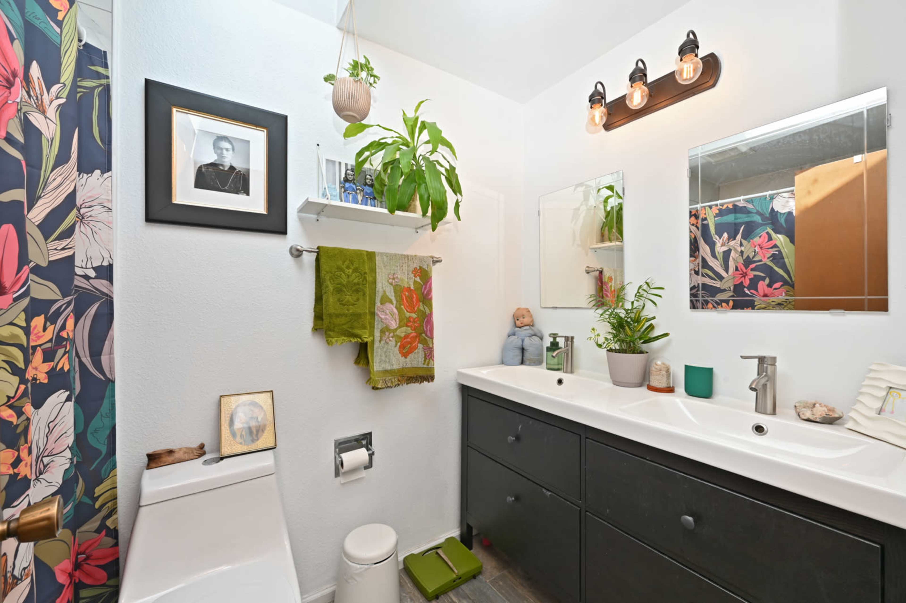 The image shows a bright bathroom featuring a dark vanity with a white countertop, a mirror, shelves with decorative plants, and floral-patterned decor.