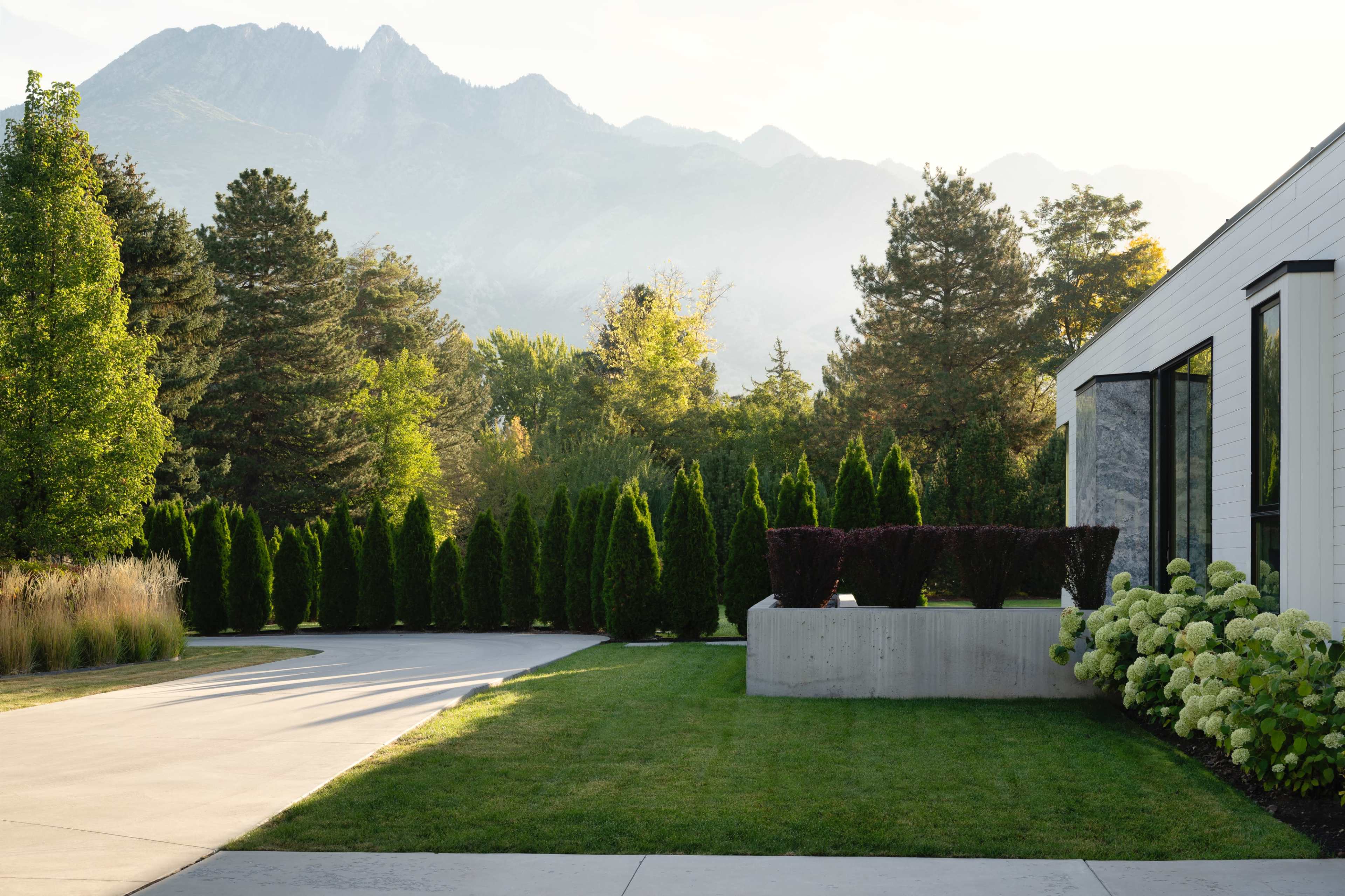 A modern house with large windows is situated next to a neatly landscaped yard featuring shrubs, a concrete planter, and a backdrop of mountains and trees.