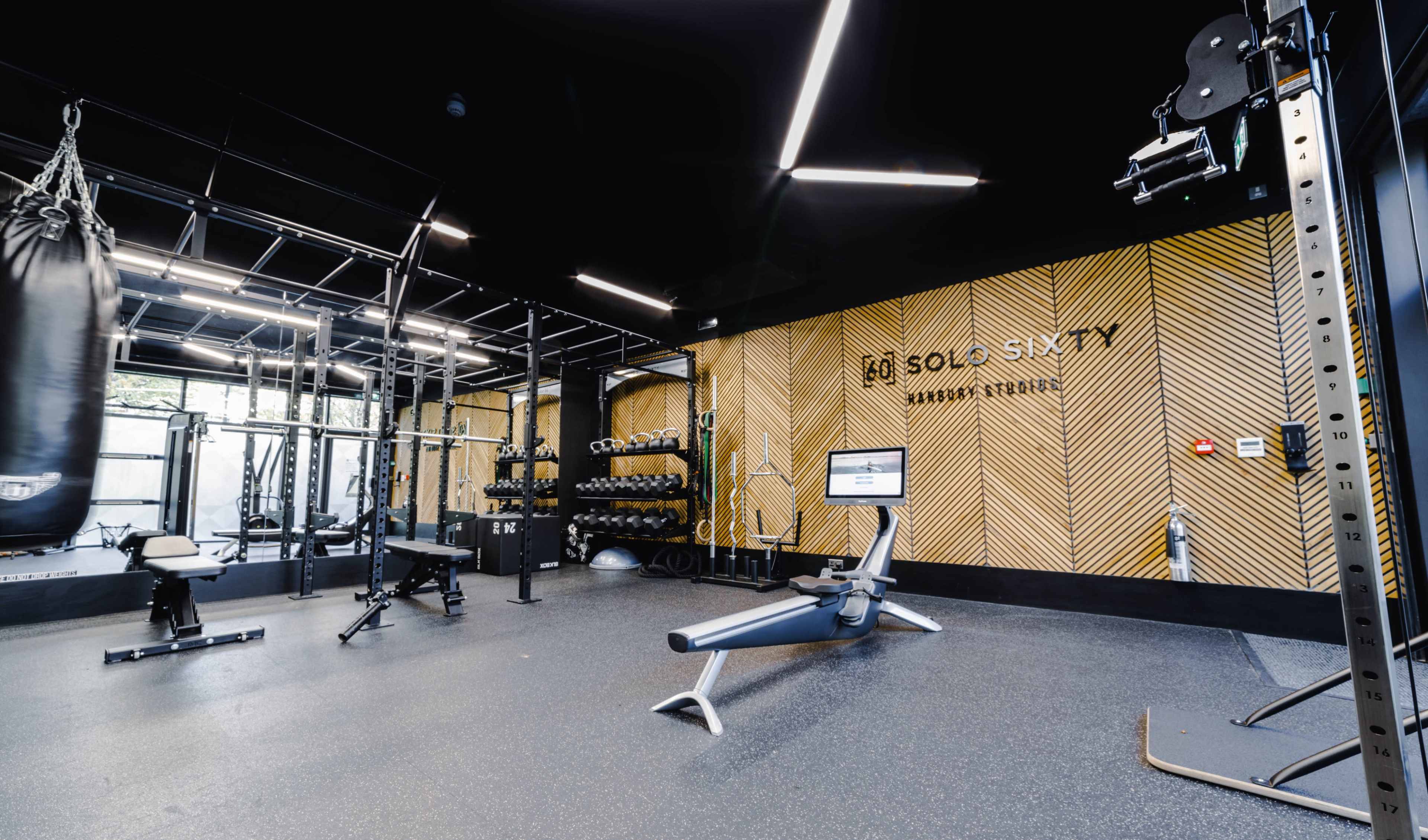 The image depicts a modern gym interior featuring various exercise equipment, including weights, a rowing machine, and a punching bag, against a backdrop of patterned wall design.