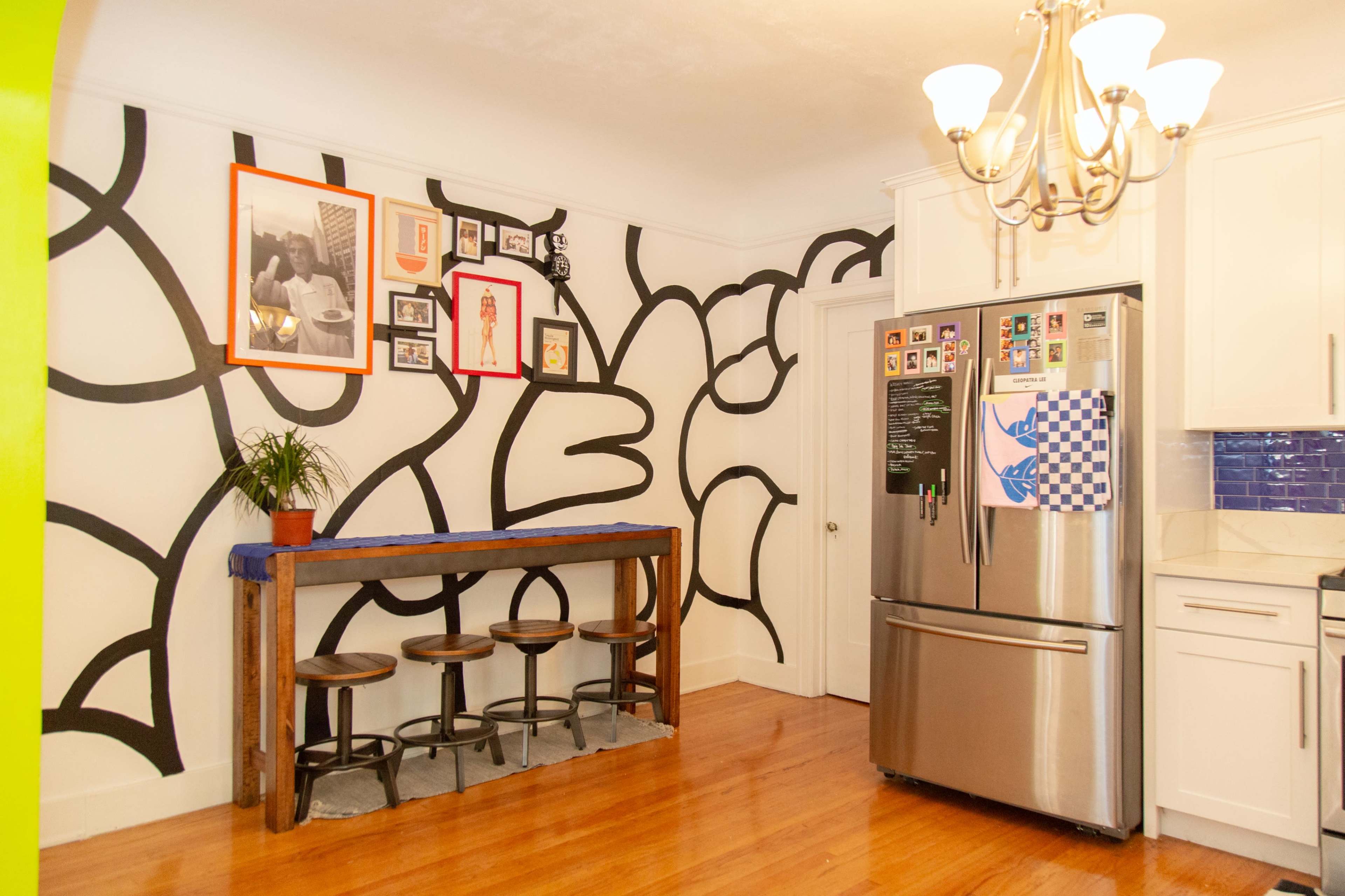 The kitchen features a wall with abstract black lines, three wooden bar stools, and a stainless steel refrigerator next to white cabinets.