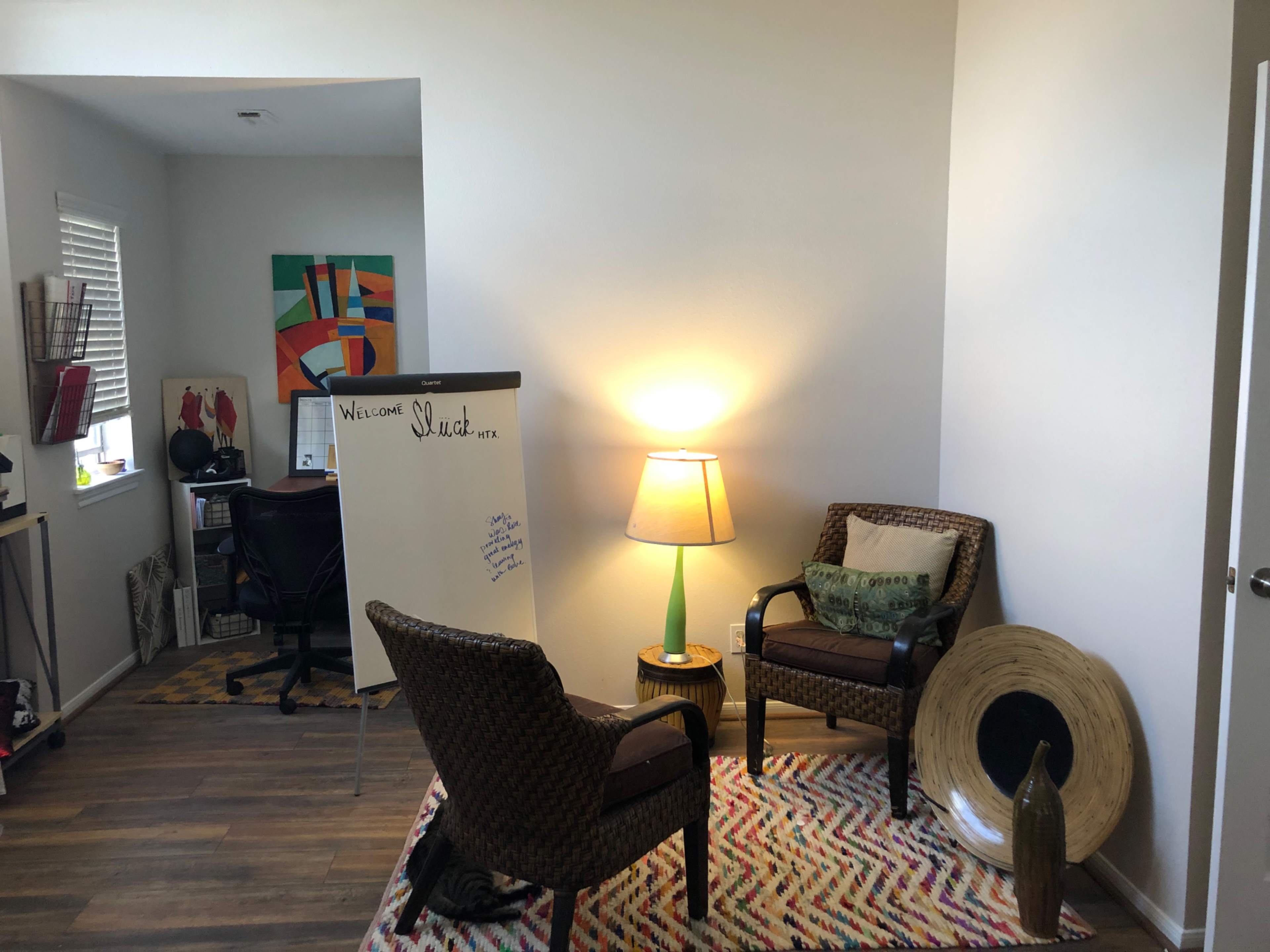 A well-lit room featuring two woven chairs, a lamp, a whiteboard, and a desk area with artwork on the walls.