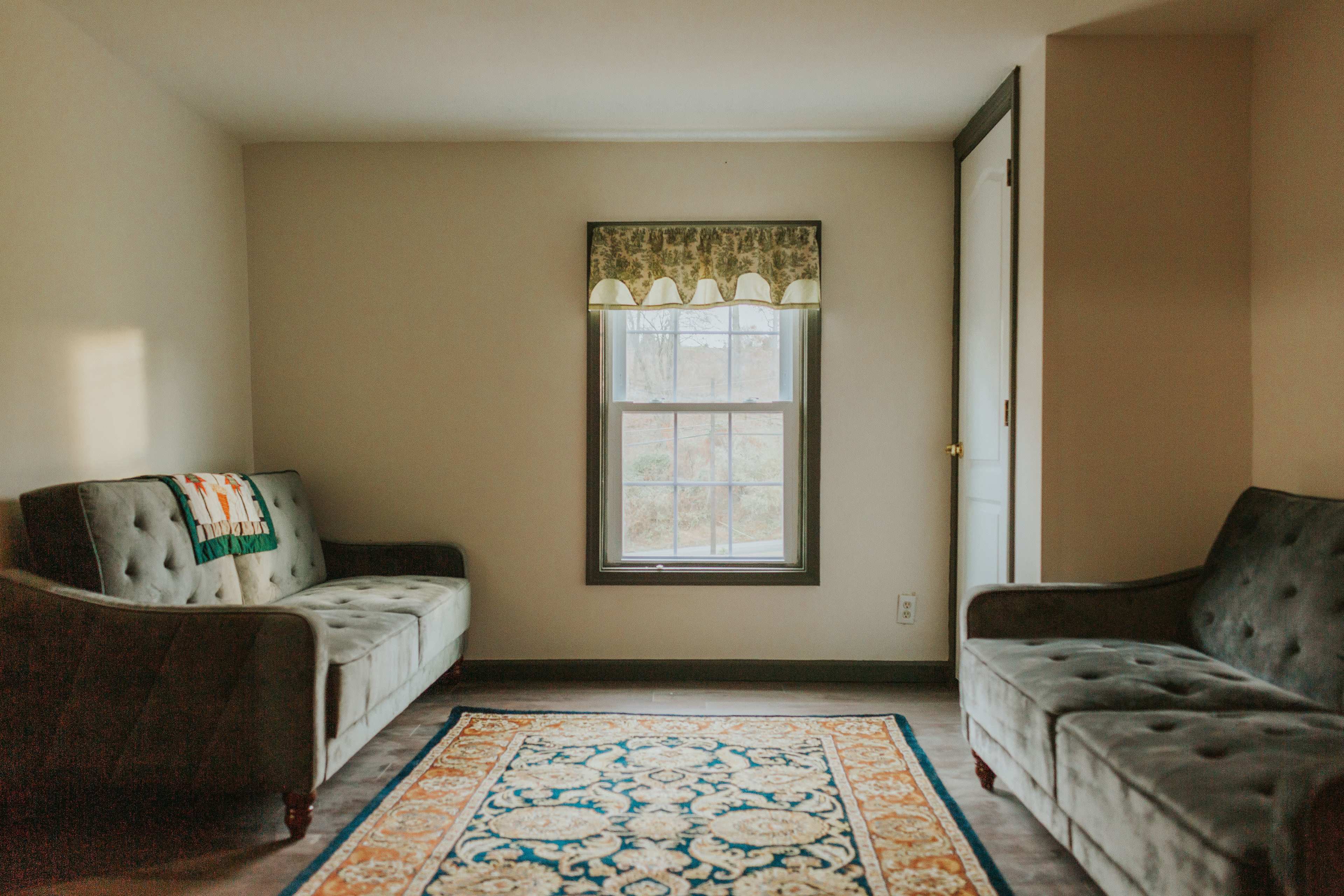 A simple living room with two sofas facing each other, a large area rug on the floor, and a window with patterned curtains.