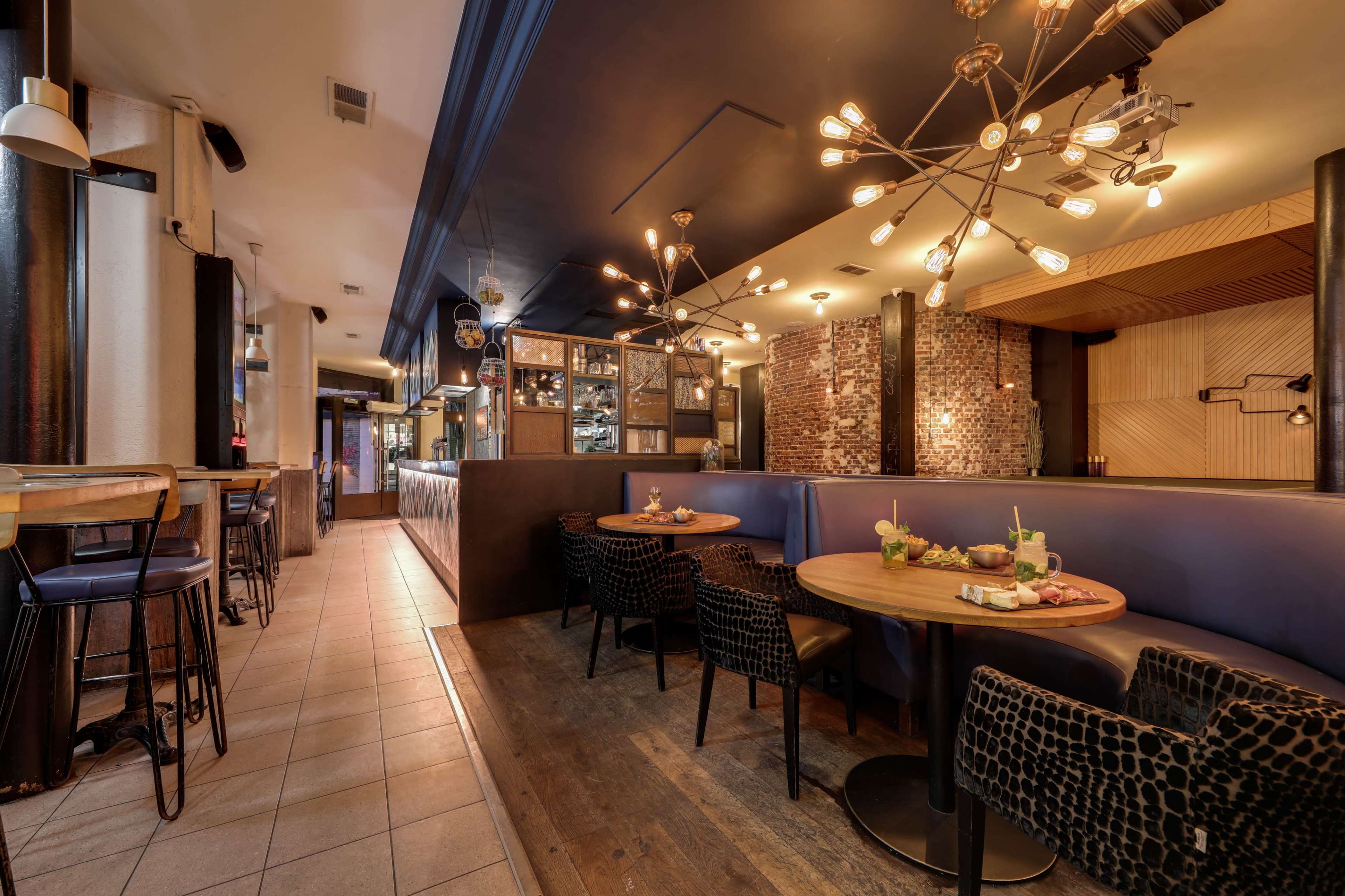 The image shows a modern restaurant interior featuring wooden tables, black chairs, and decorative lighting, with a partial view of the bar area in the background.