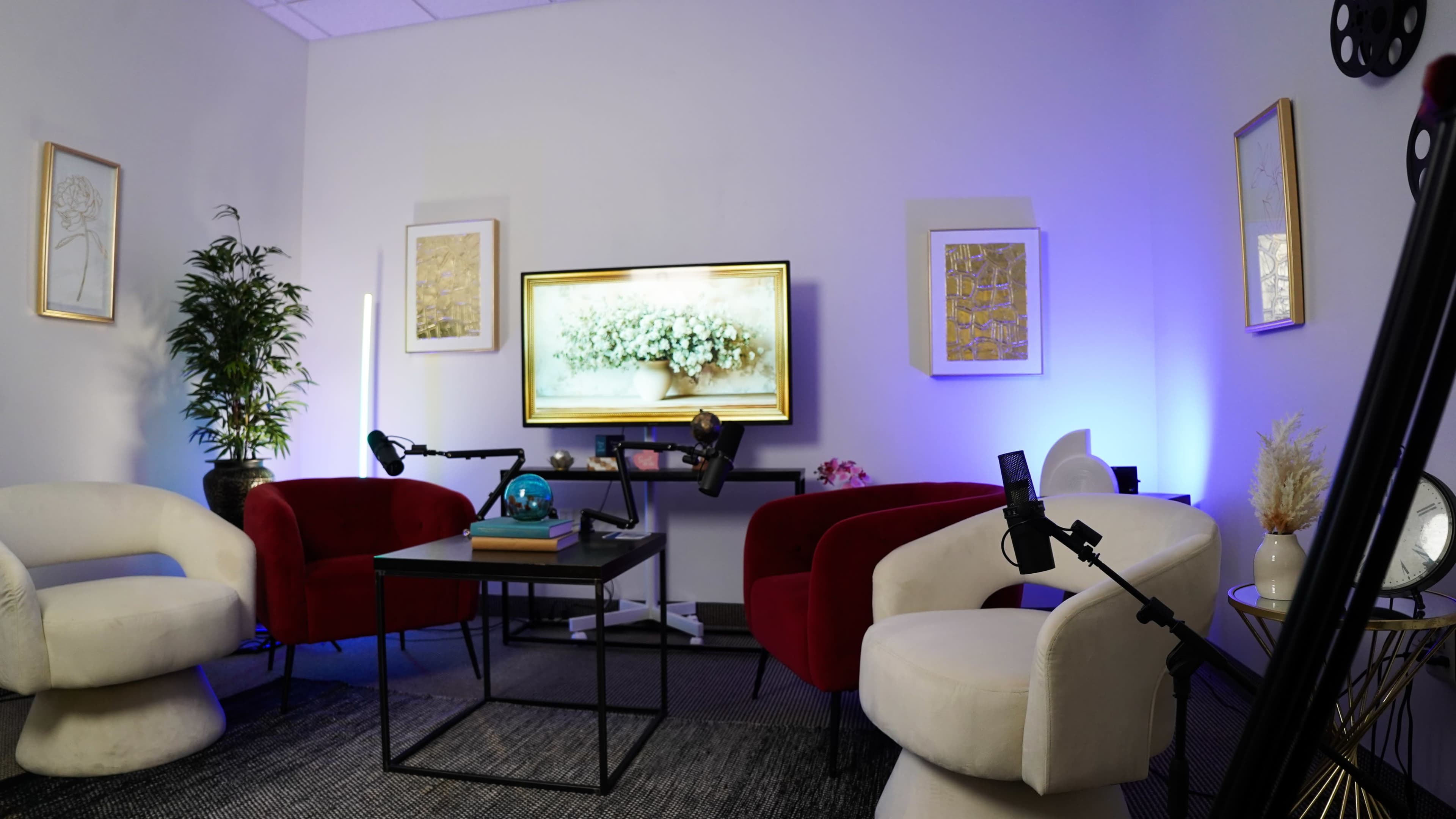 The scene features a cozy lounge area with two red chairs and one white chair, a black table in the center, and a television displaying a floral image on a wall, accompanied by decorative artwork.