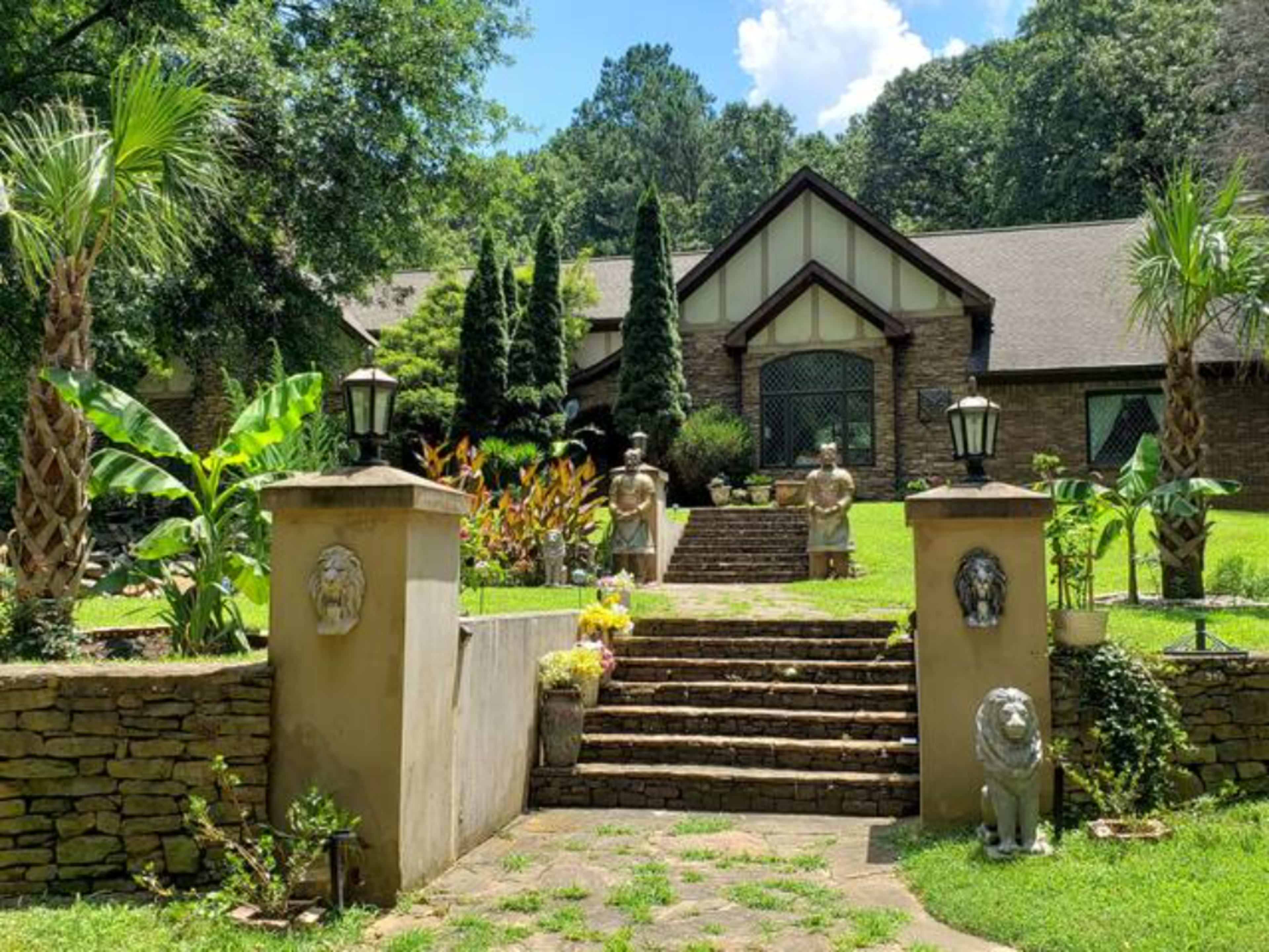 A stone pathway leading to a large house surrounded by lush greenery, palm trees, and decorative sculptures.