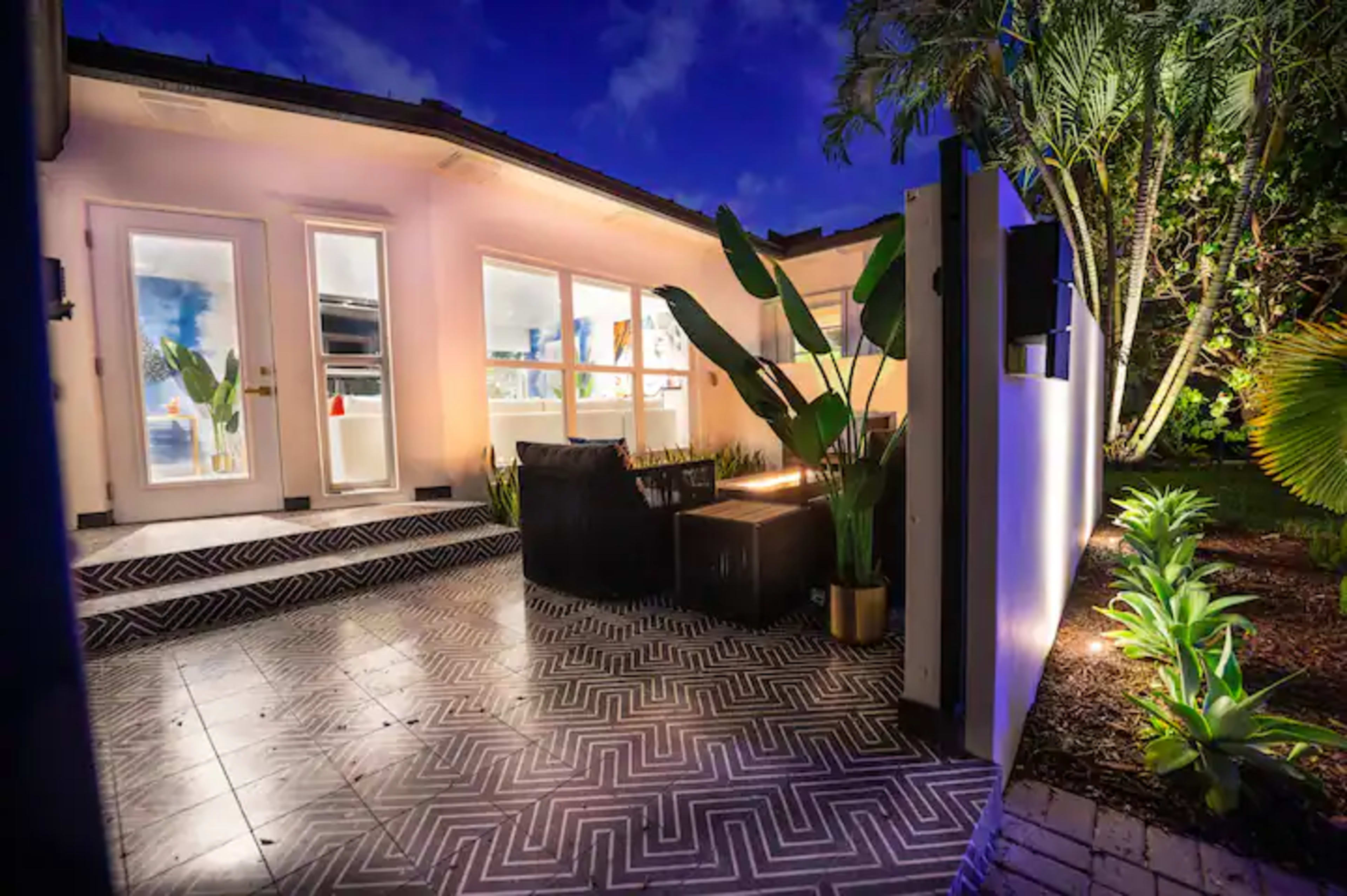 A well-lit outdoor patio with geometric tile flooring, featuring lounge seating and surrounding tropical plants.