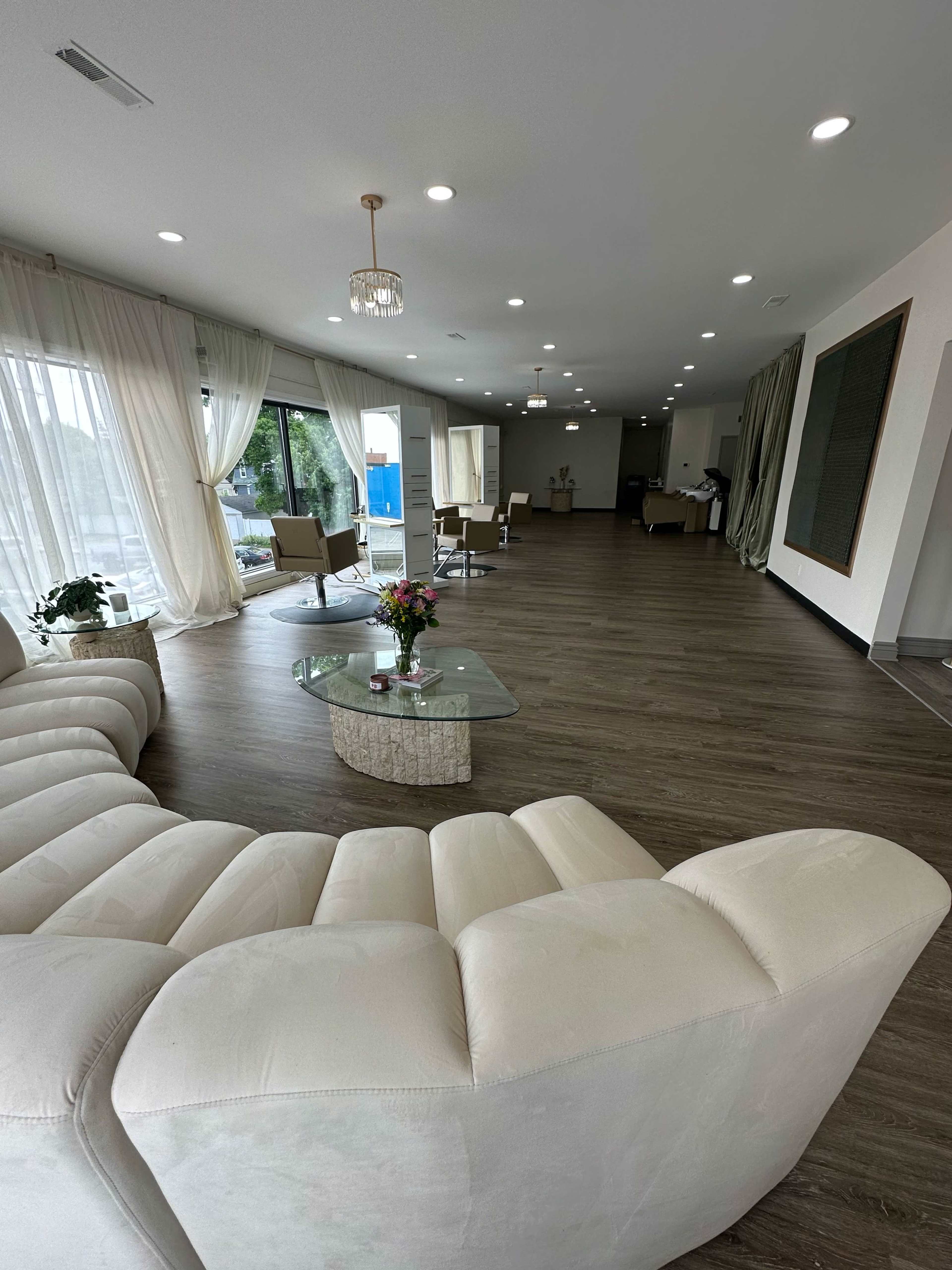 A spacious, modern interior featuring a light-colored sofa, a glass coffee table, and large windows draped with sheer curtains.