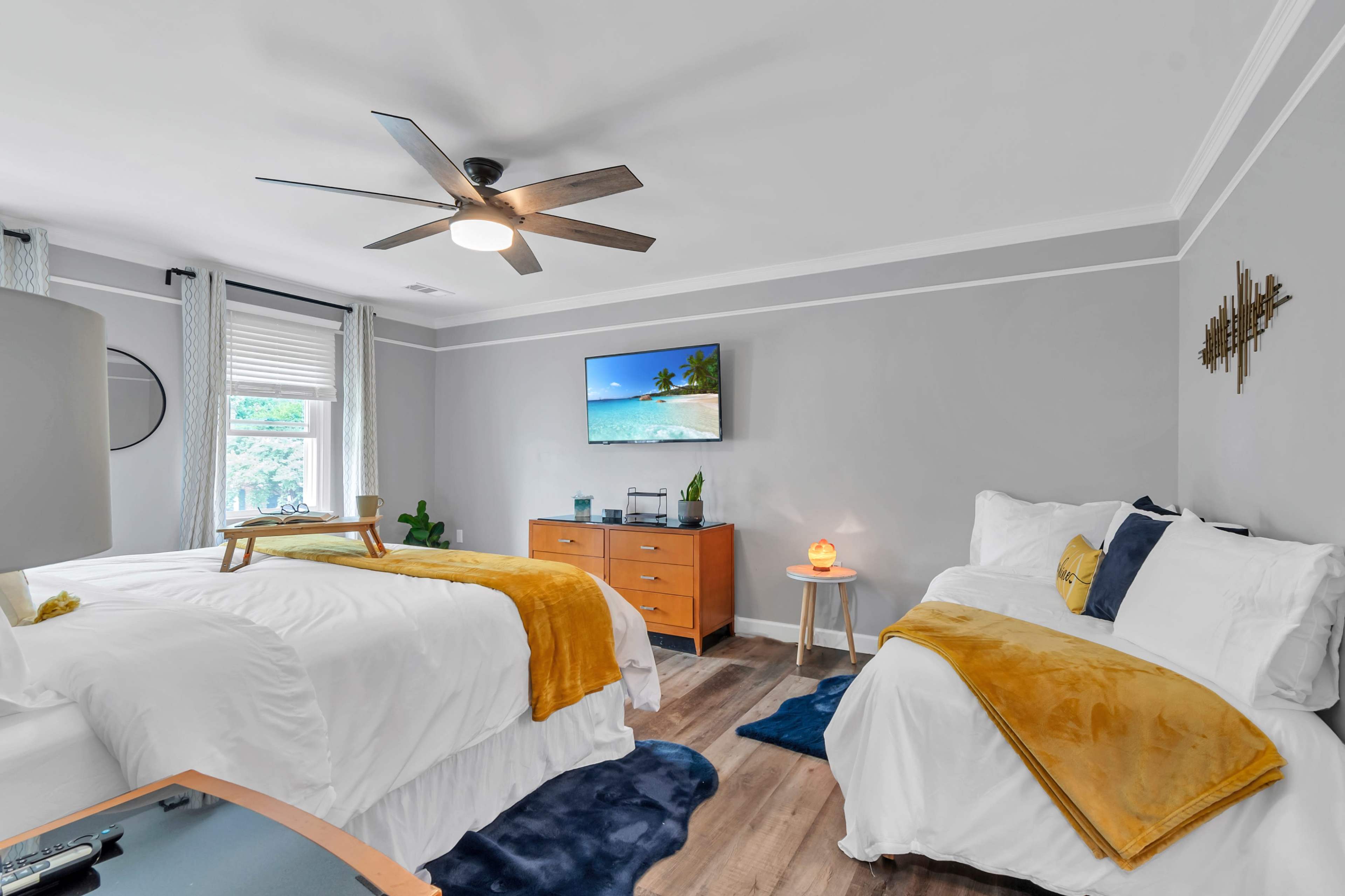 A well-decorated bedroom with two beds, a television mounted on the wall, and a dresser, featuring neutral colors and cozy accents.