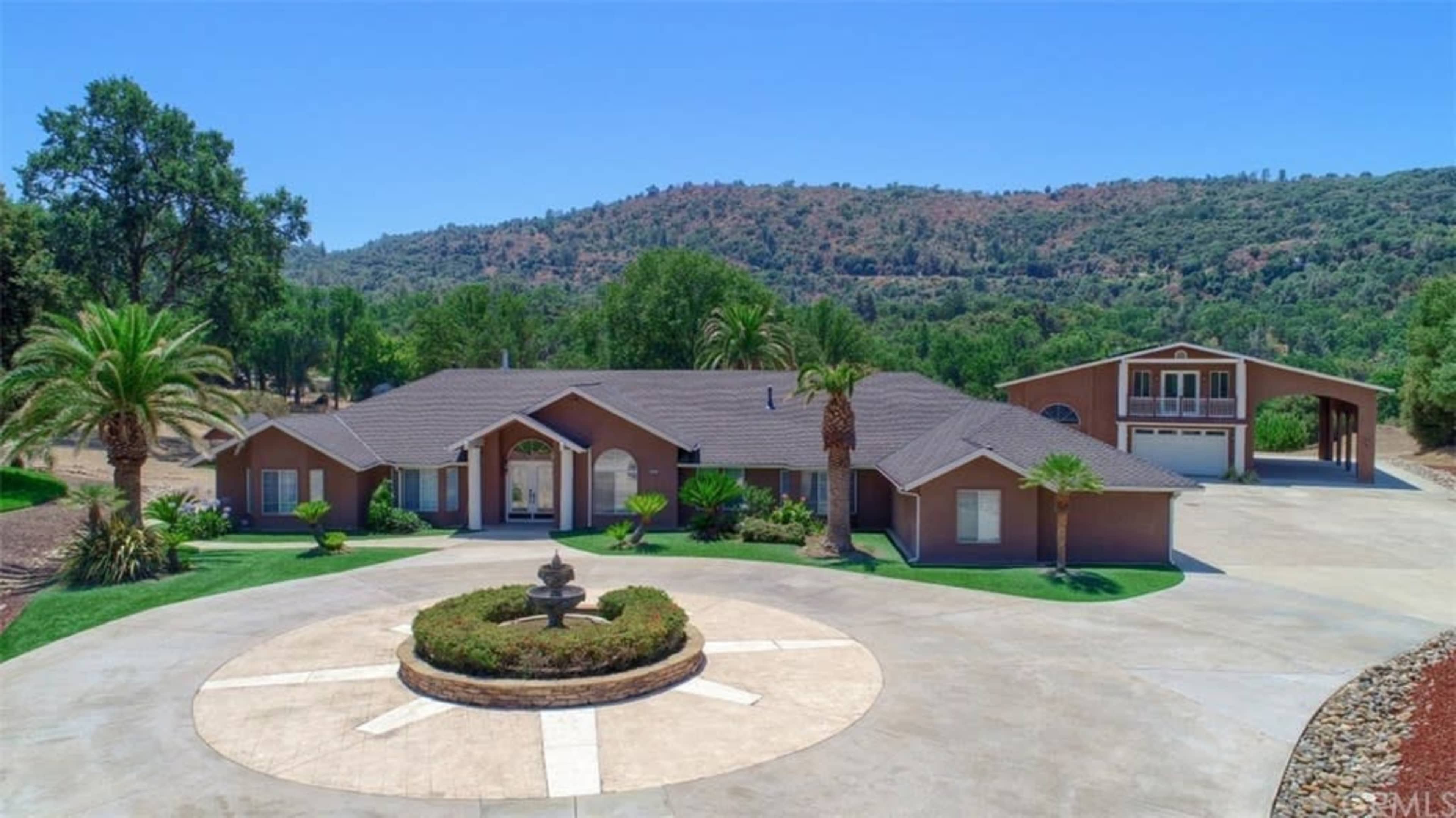 A spacious single-story home with a circular driveway and a fountain is surrounded by palm trees and hillside greenery.