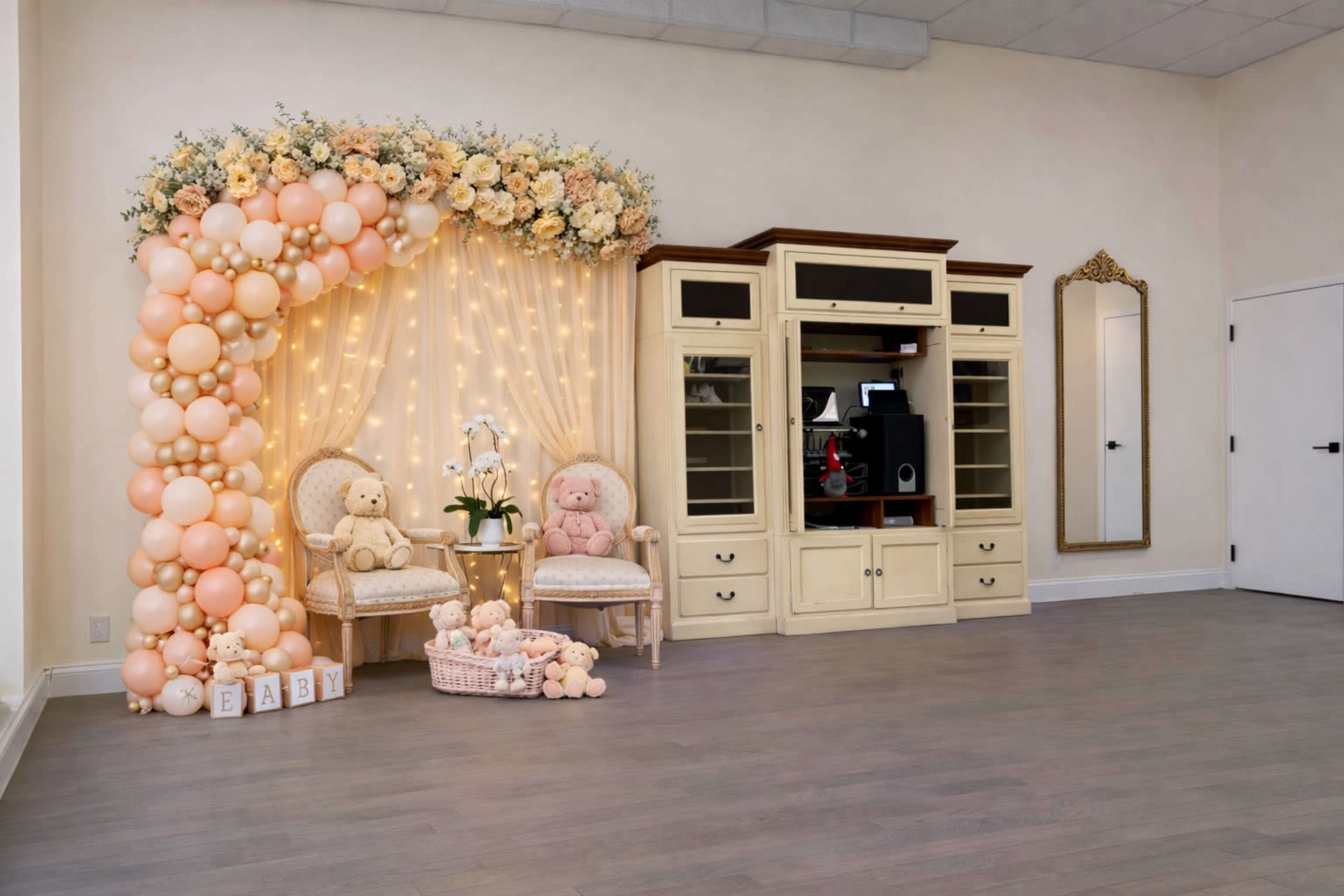 The image shows a room decorated with a balloon arch and floral arrangement, featuring two teddy bears seated on decorative chairs next to a wooden cabinet and a mirror.