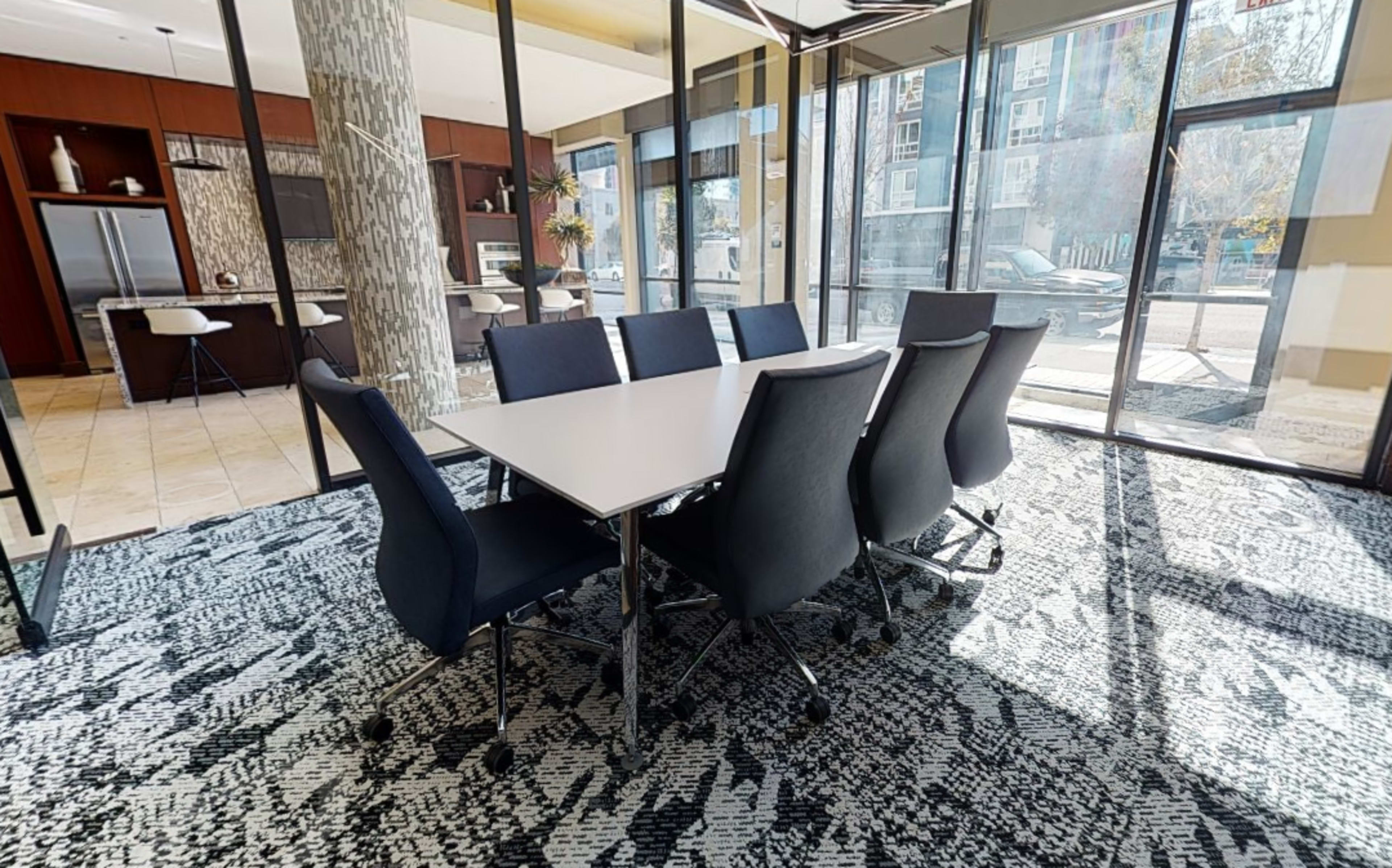 A modern conference room features a large white table surrounded by eight dark chairs, with glass walls allowing natural light and a view of the outside.