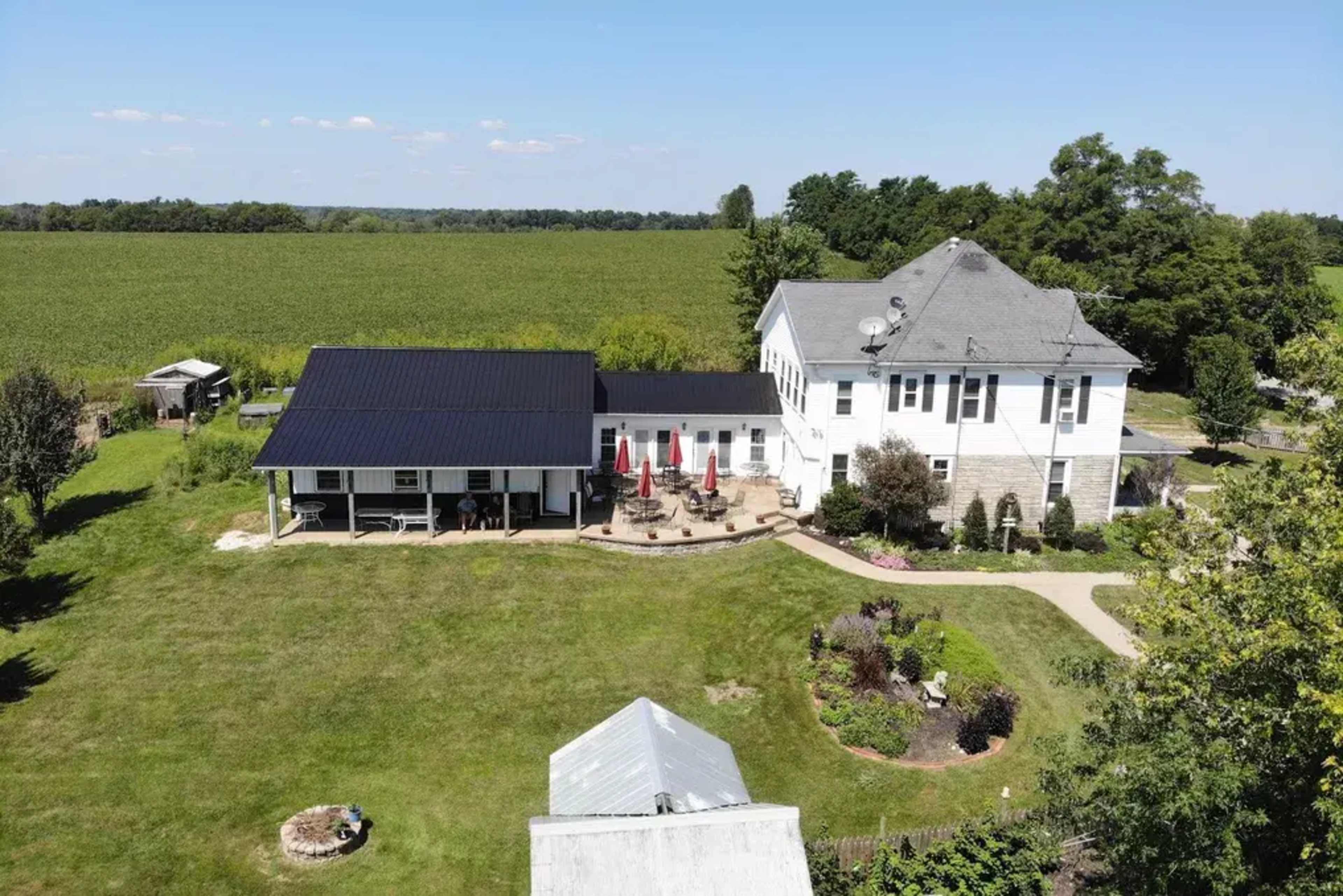 A two-story house with a gray exterior and a black roof is adjacent to a single-story covered patio area with red umbrellas, surrounded by a grassy yard and fields in the background.