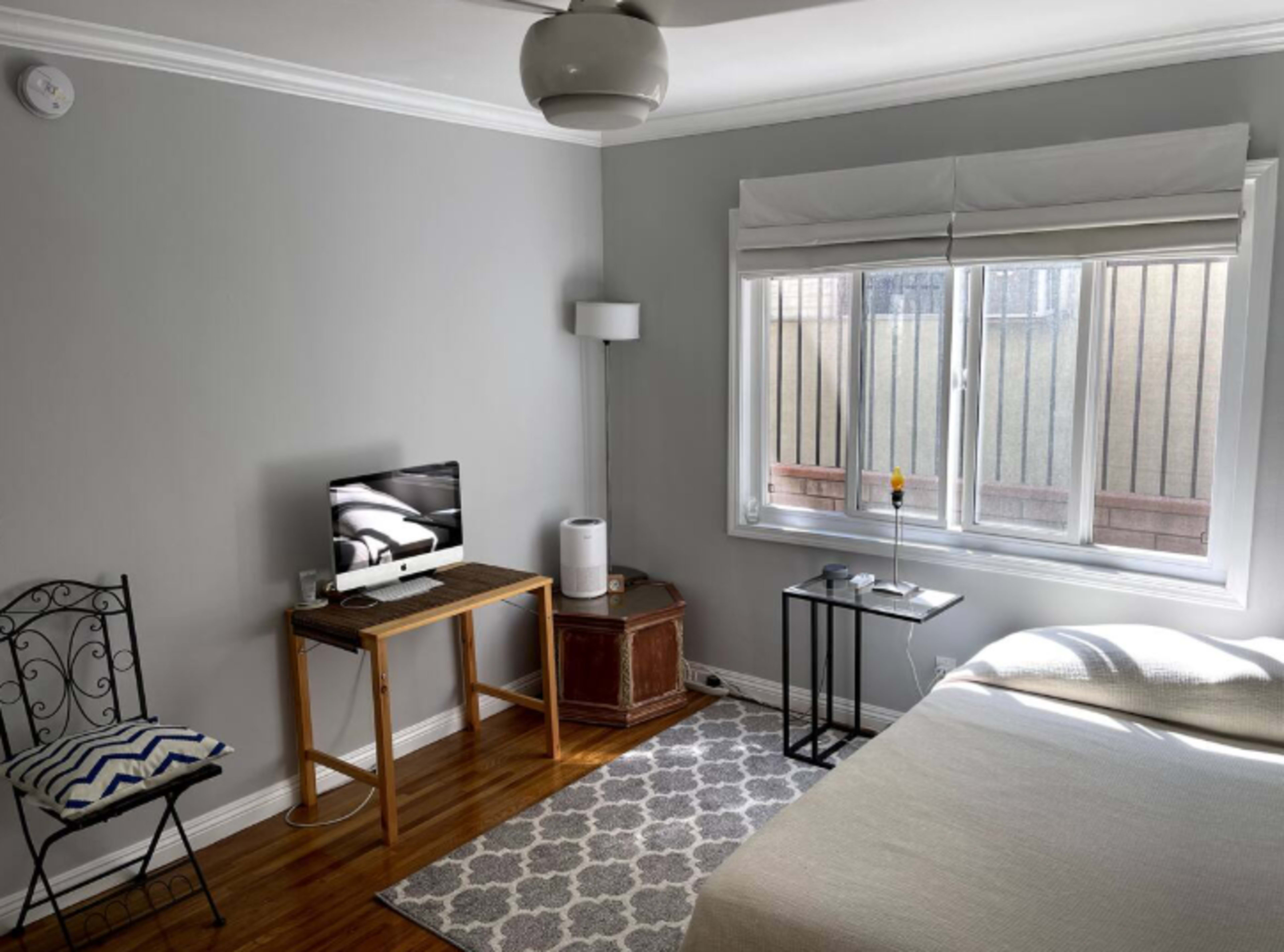 The image shows a minimalistic bedroom featuring a bed, a small table, a chair, and a window with light filtering through.