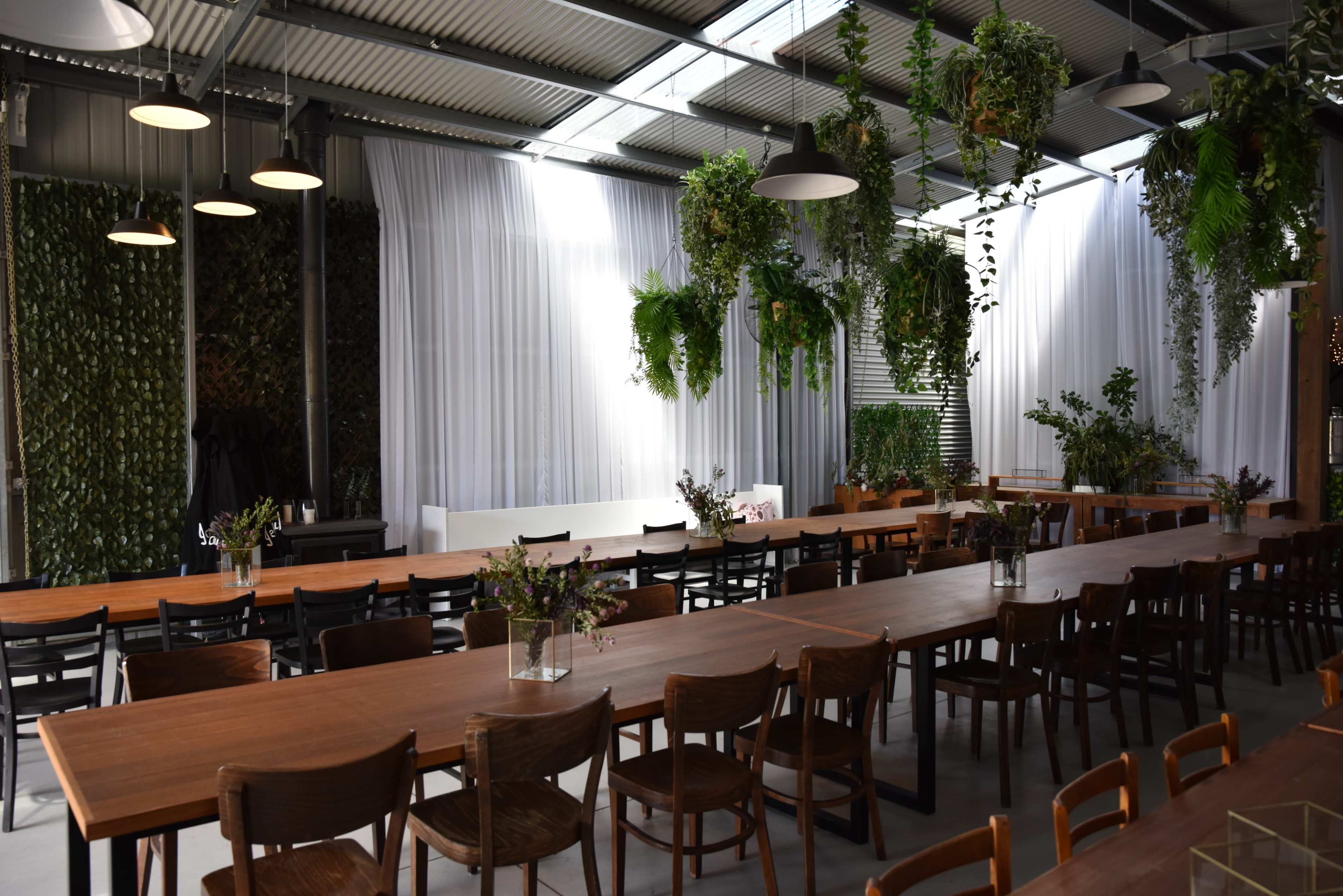 The image shows a modern dining area with multiple long wooden tables, surrounded by plants and greenery, illuminated by natural light filtering through large windows.