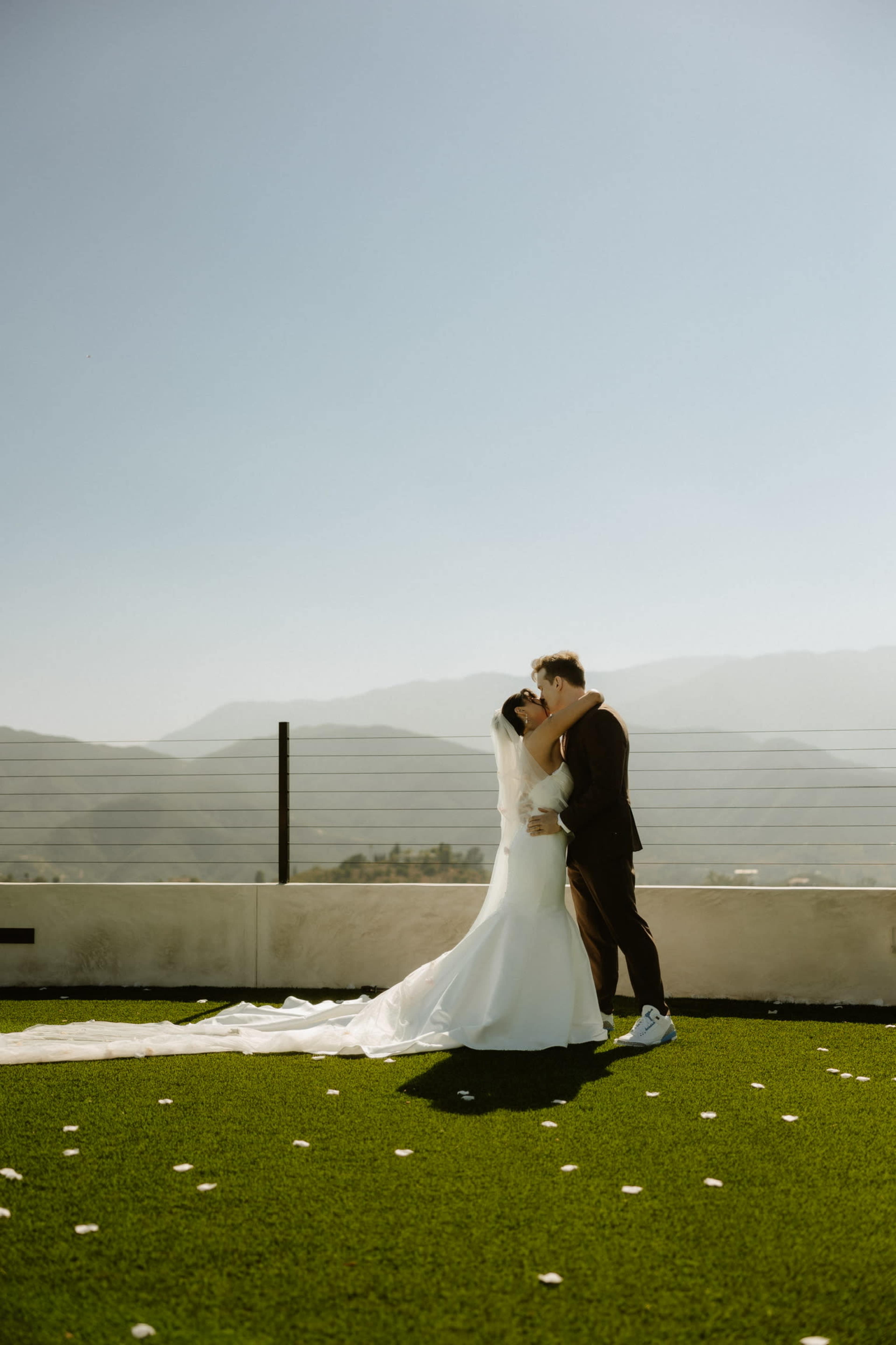 A couple shares a kiss on a rooftop, surrounded by mountains and a green carpet of artificial grass, with rose petals scattered on the ground.