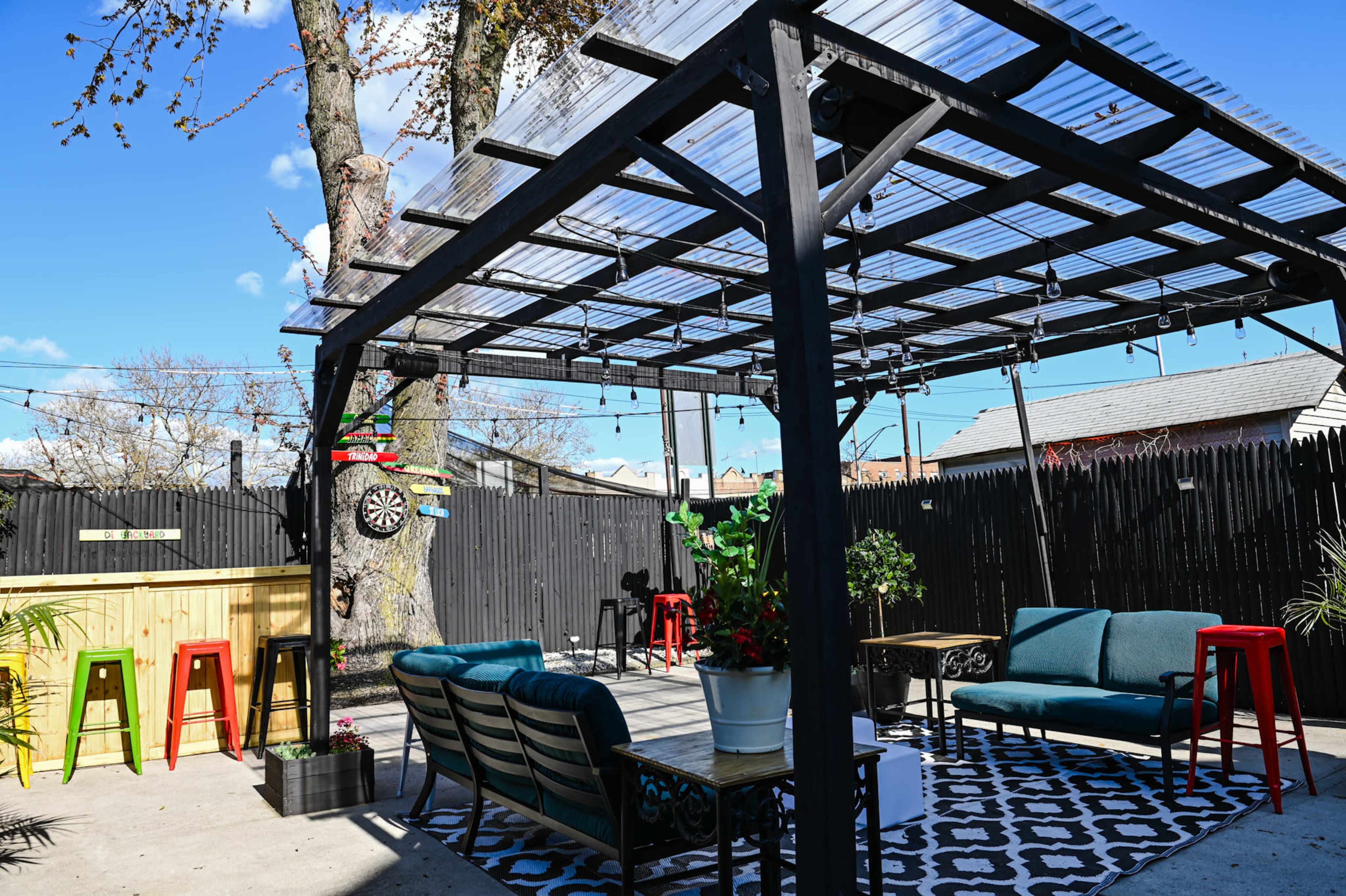 A patio area features a pergola with string lights, colorful seating, and a dartboard, surrounded by a wooden fence.