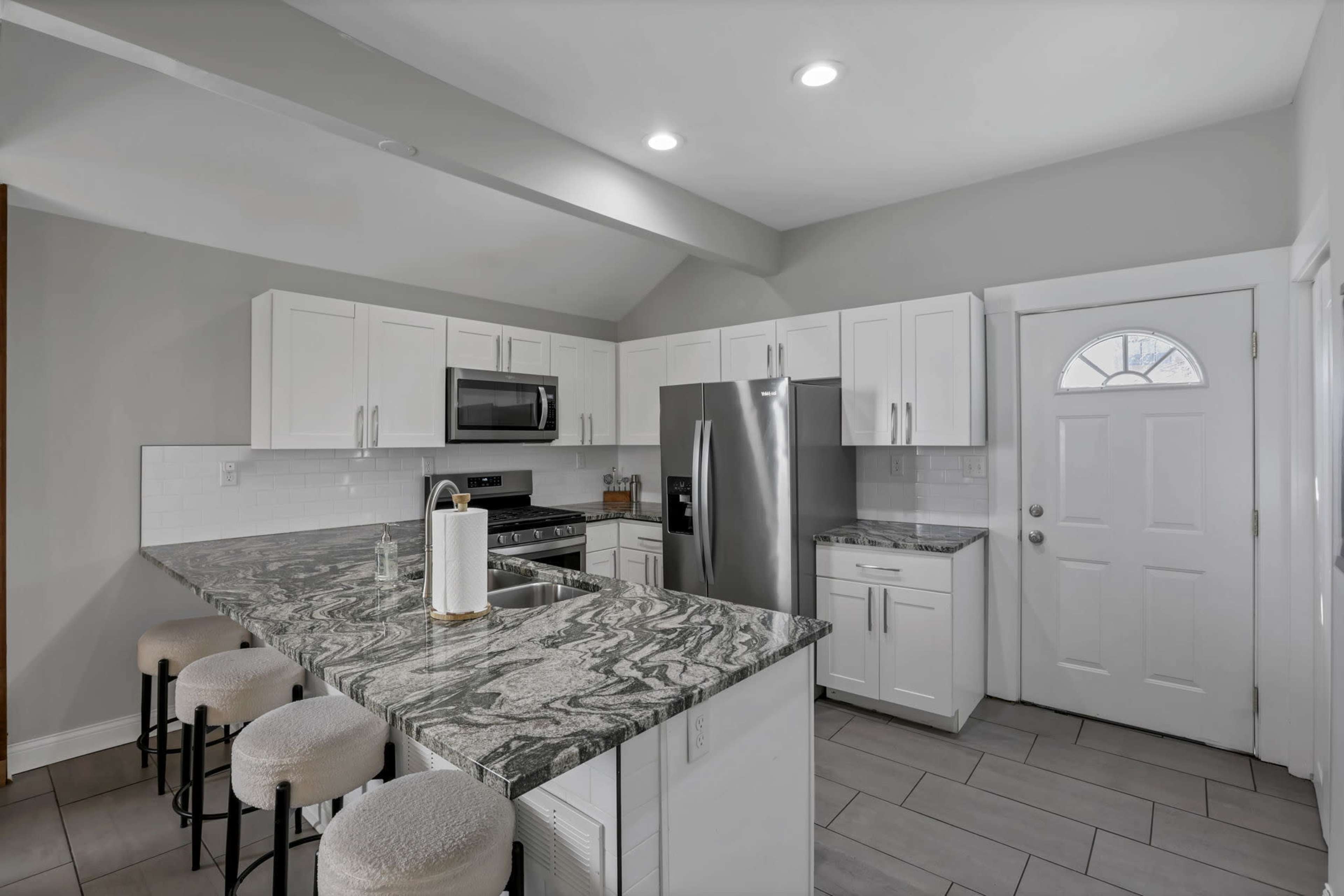 The image shows a modern kitchen with white cabinetry, stainless steel appliances, and a marble-patterned countertop with bar stools.