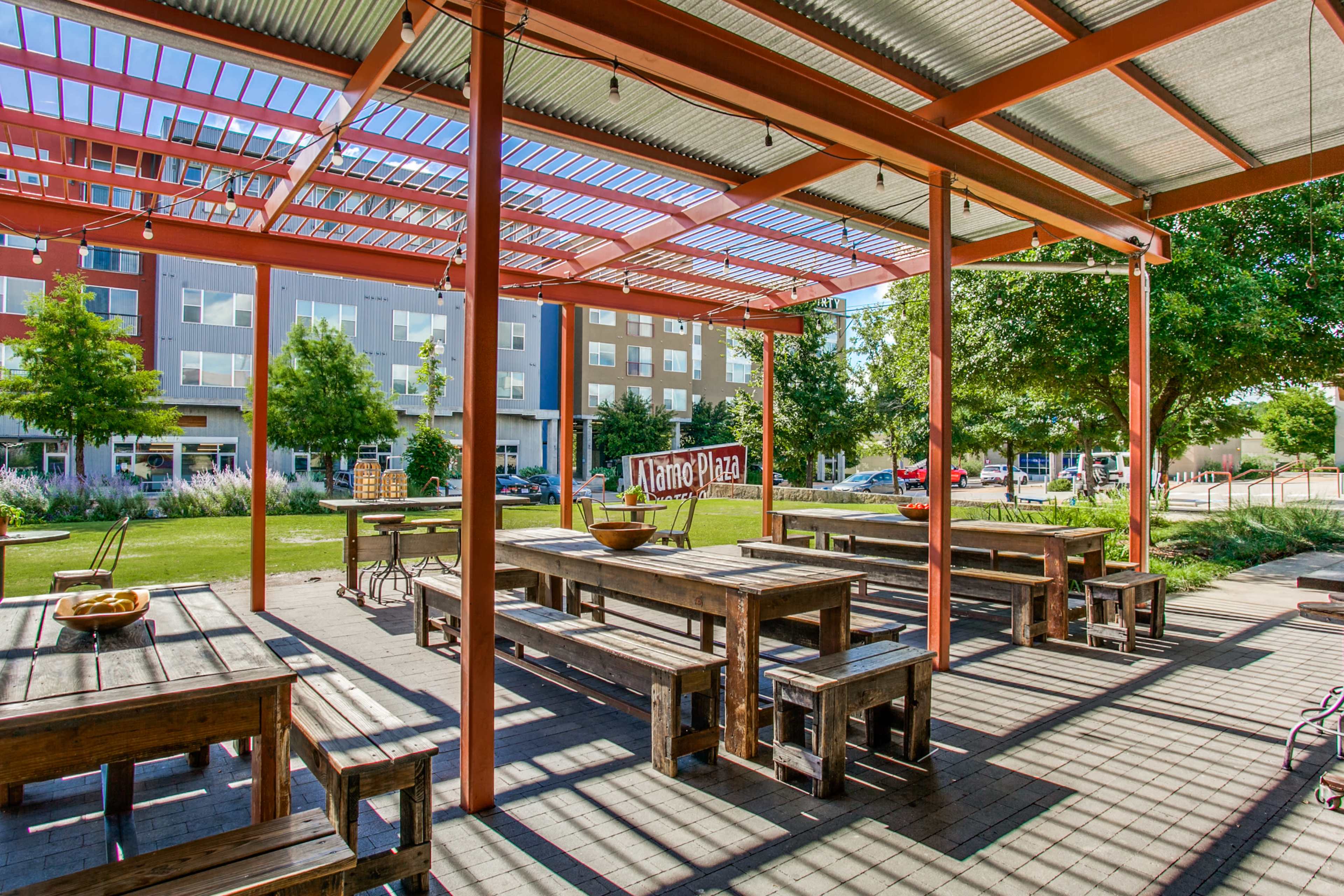 An outdoor dining area features wooden tables and benches under a pergola, with green grass and modern buildings in the background.