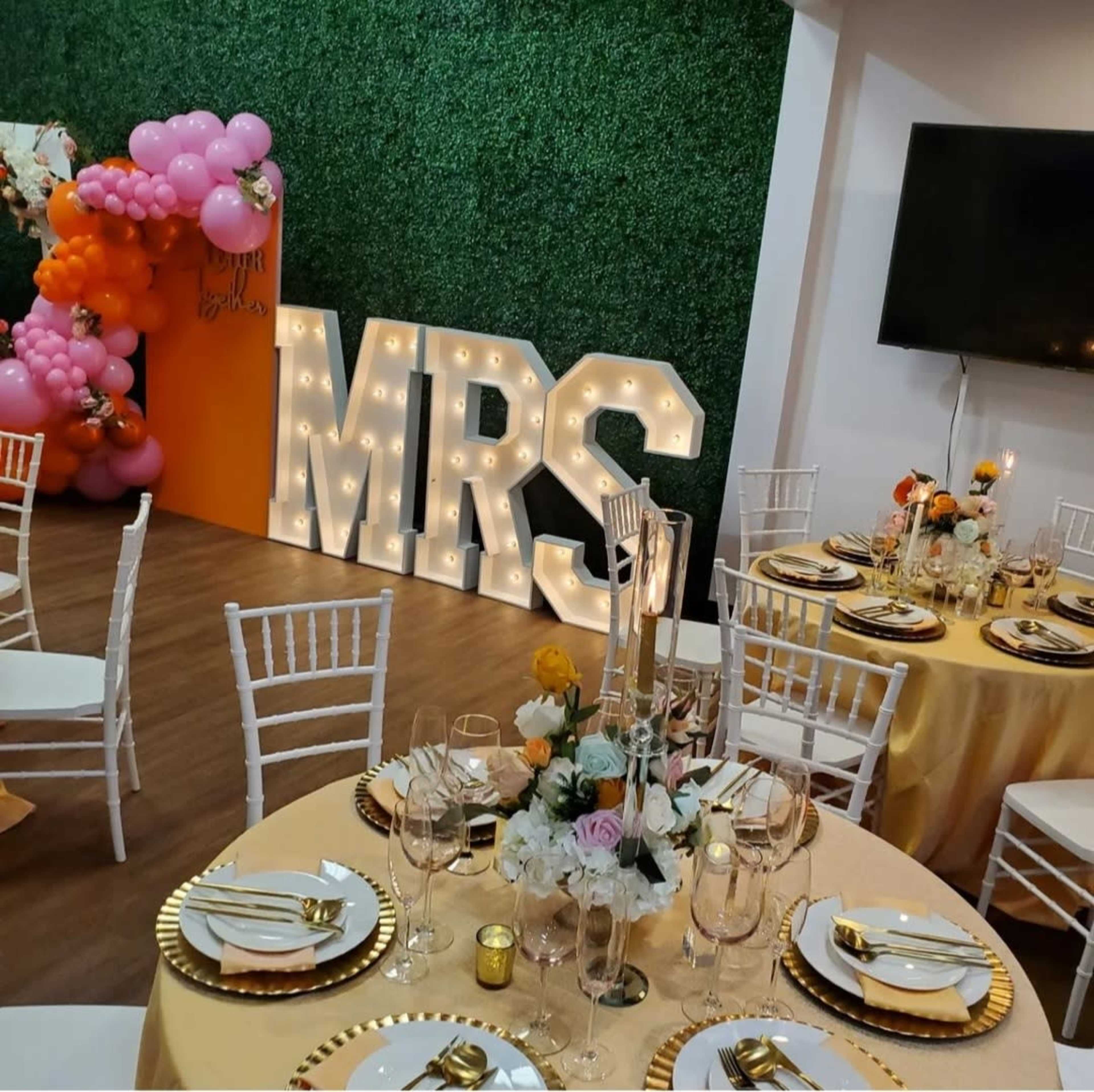 A decorated event space with a large "MRS" sign, floral arrangements on elegantly set tables, and a backdrop of greenery and balloons.