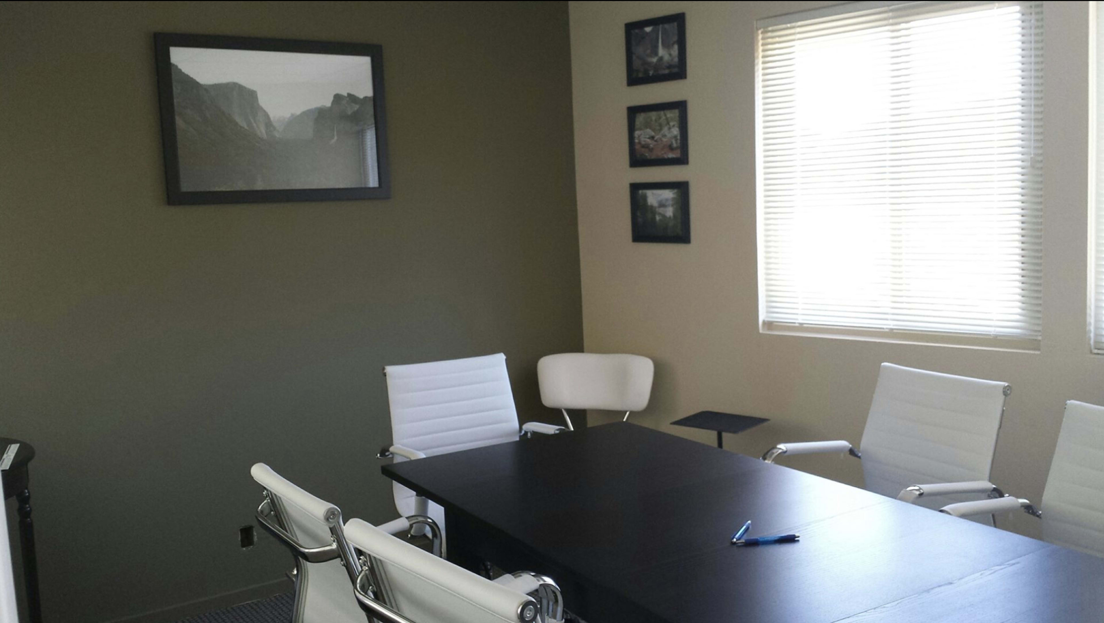 The image shows a modern conference room with a dark table surrounded by white chairs, framed pictures on the walls, and natural light coming through a window.