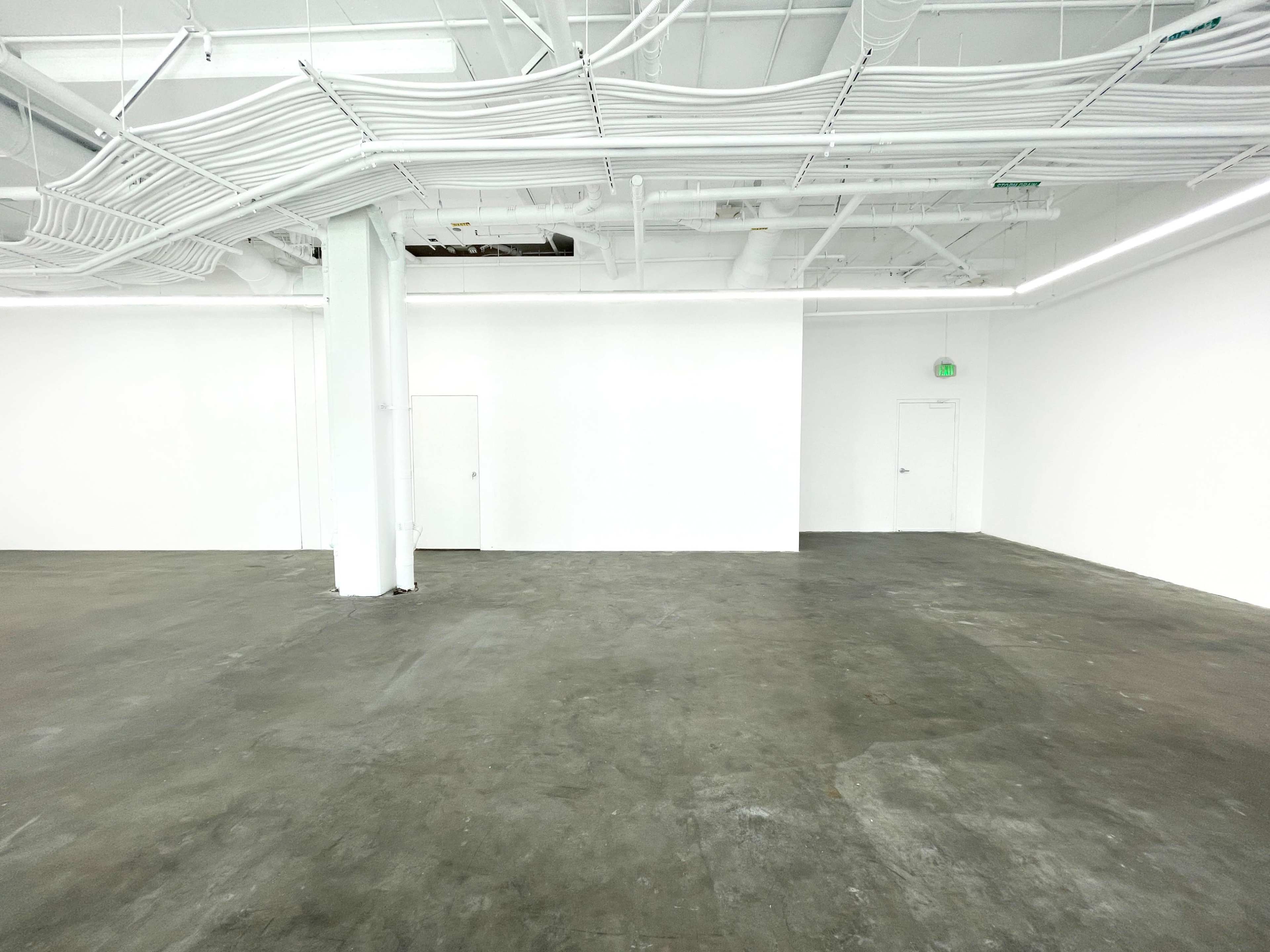 The image shows a spacious, empty room with white walls, a concrete floor, and visible overhead piping.