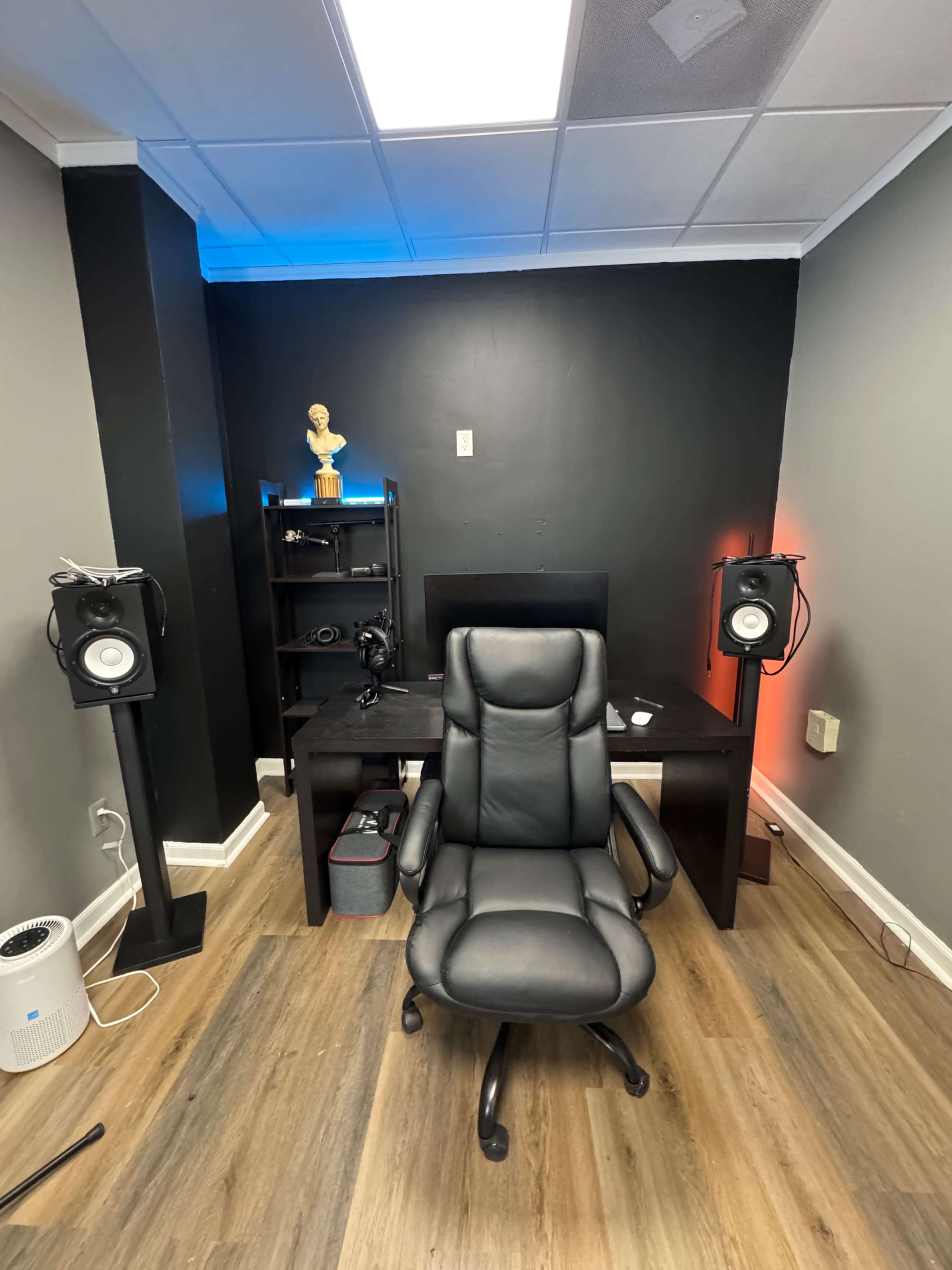 The image shows an interior room with a black desk, an office chair, speakers on stands, a small shelf, and a decorative statue, all against a dark wall.