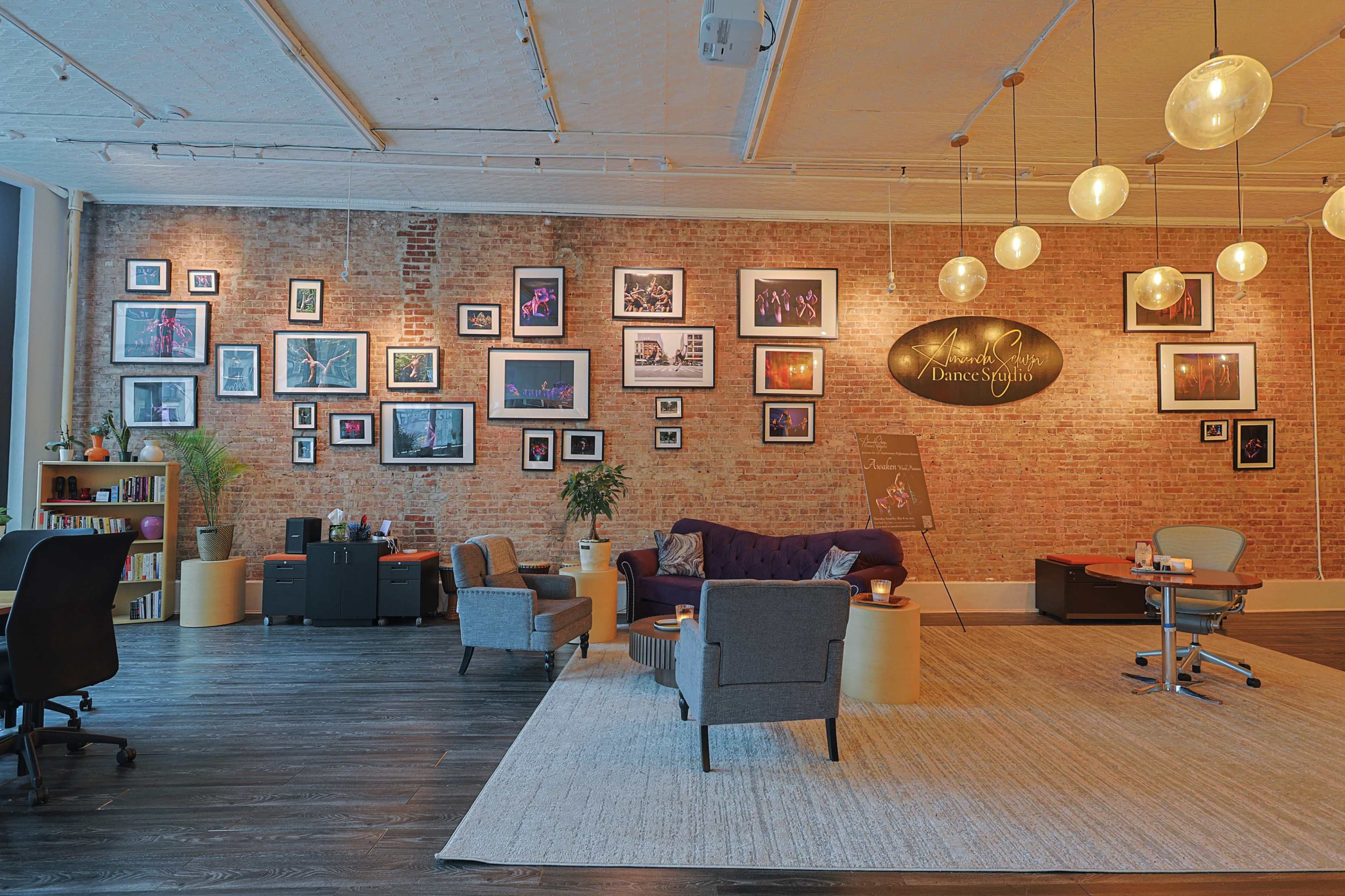 A spacious office features a brick wall adorned with framed photographs, a plush purple sofa, and several seating areas arranged on a light-colored rug.