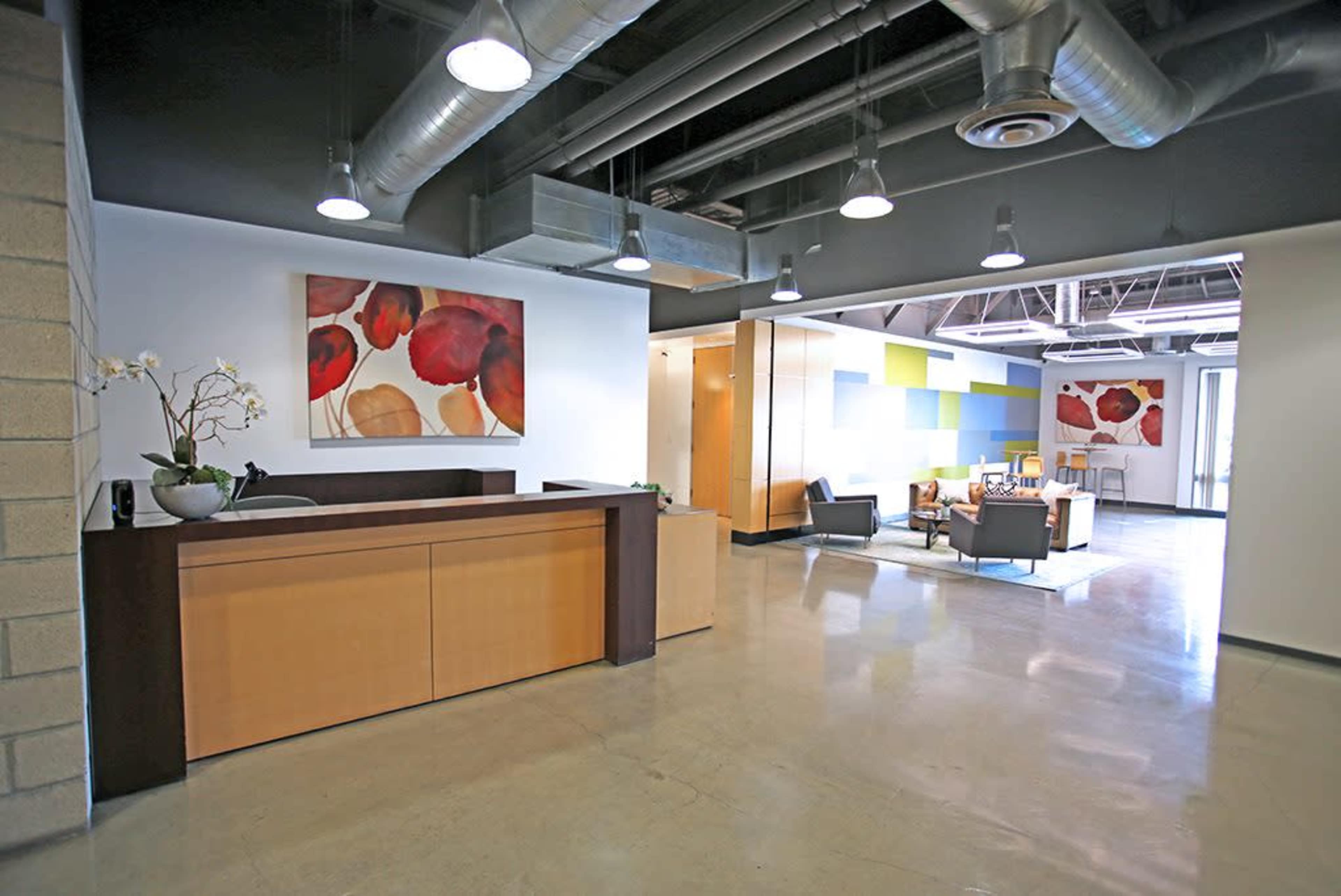 The image shows a modern office reception area with a wooden desk, colorful artwork on the walls, and a seating area in the background.