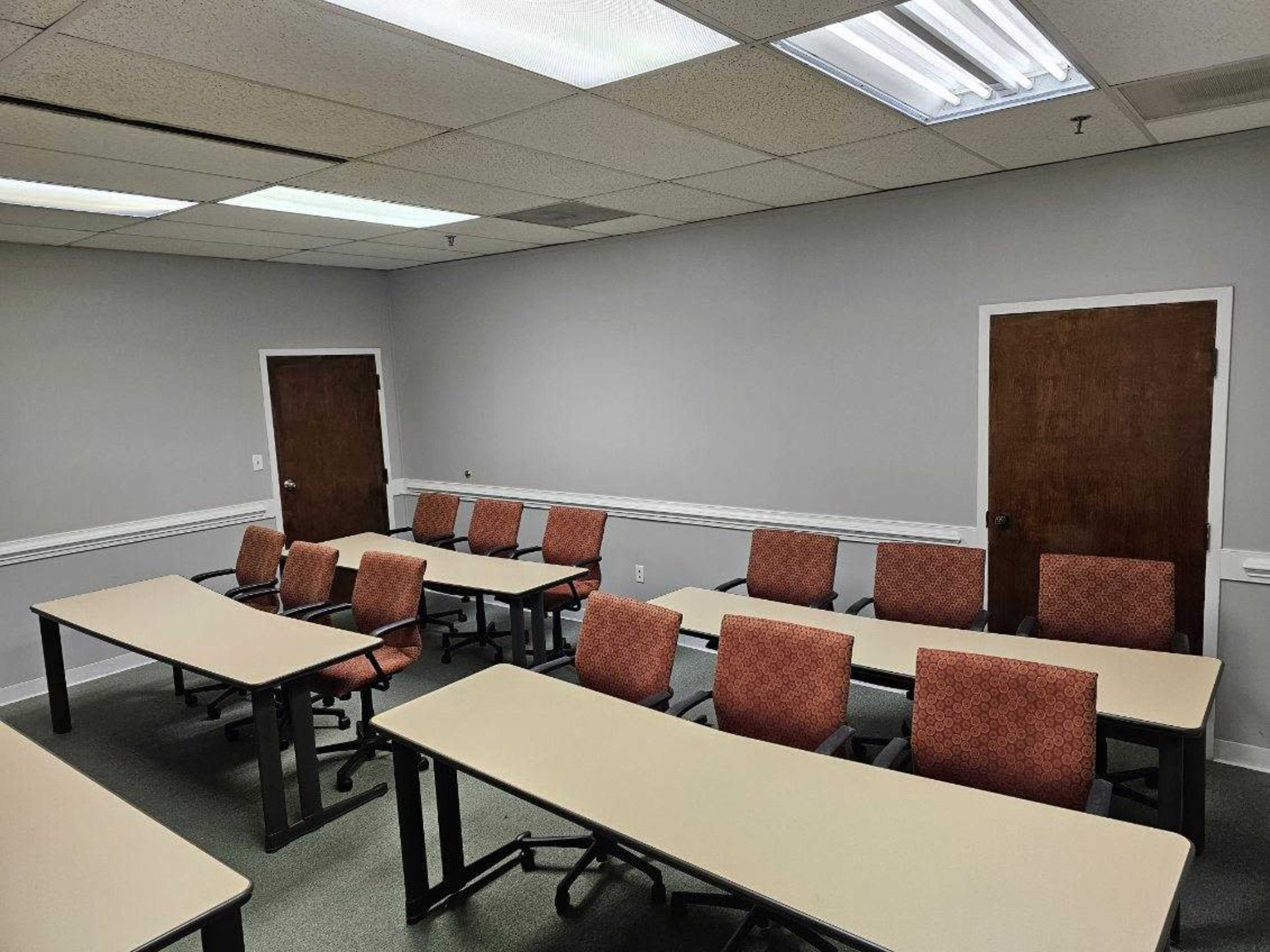 The image shows a classroom equipped with several tables and chairs arranged in a facing layout, with doors along the walls.