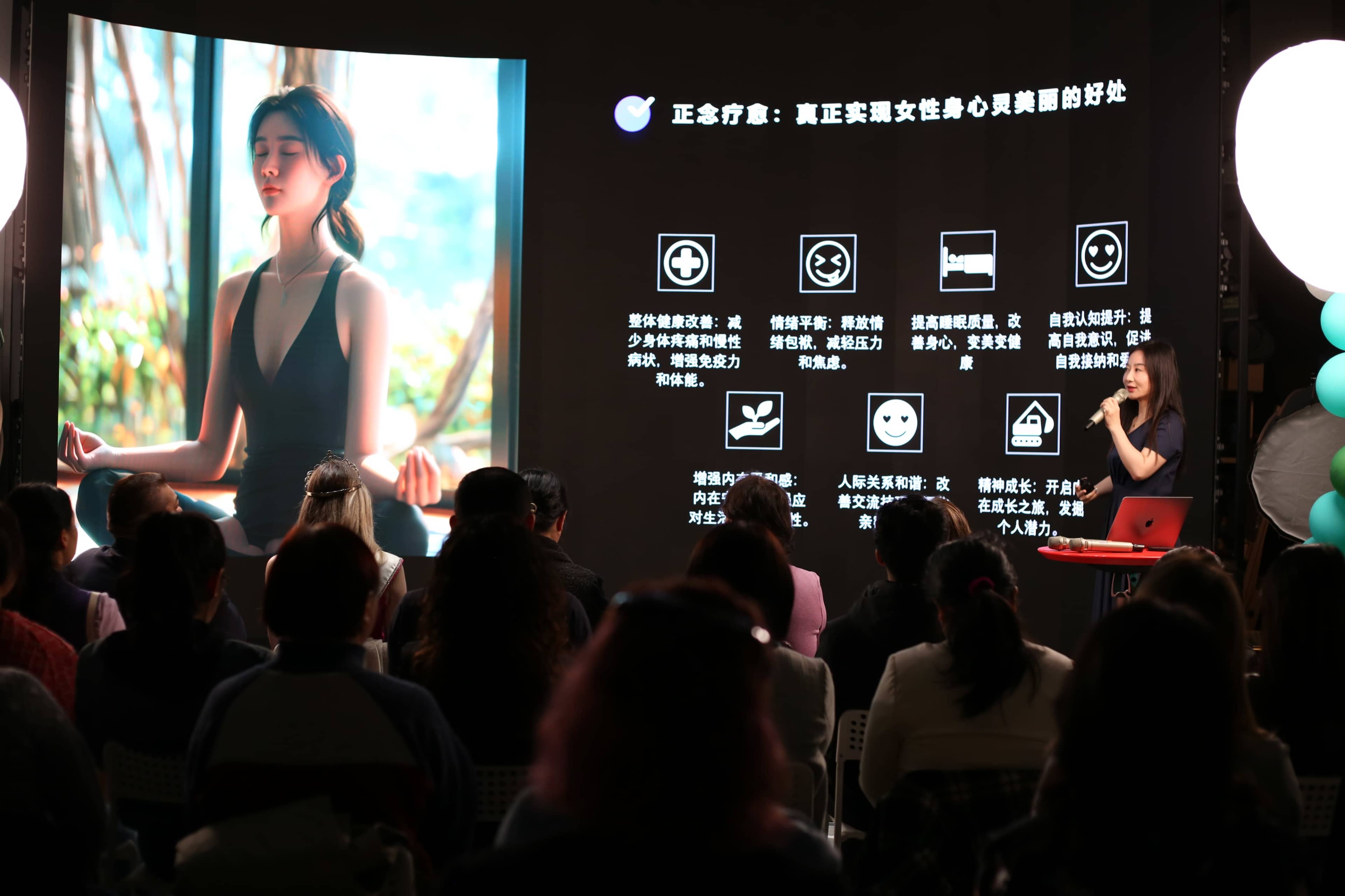 A presenter speaks to an audience while standing next to a large screen displaying information and images related to wellness.