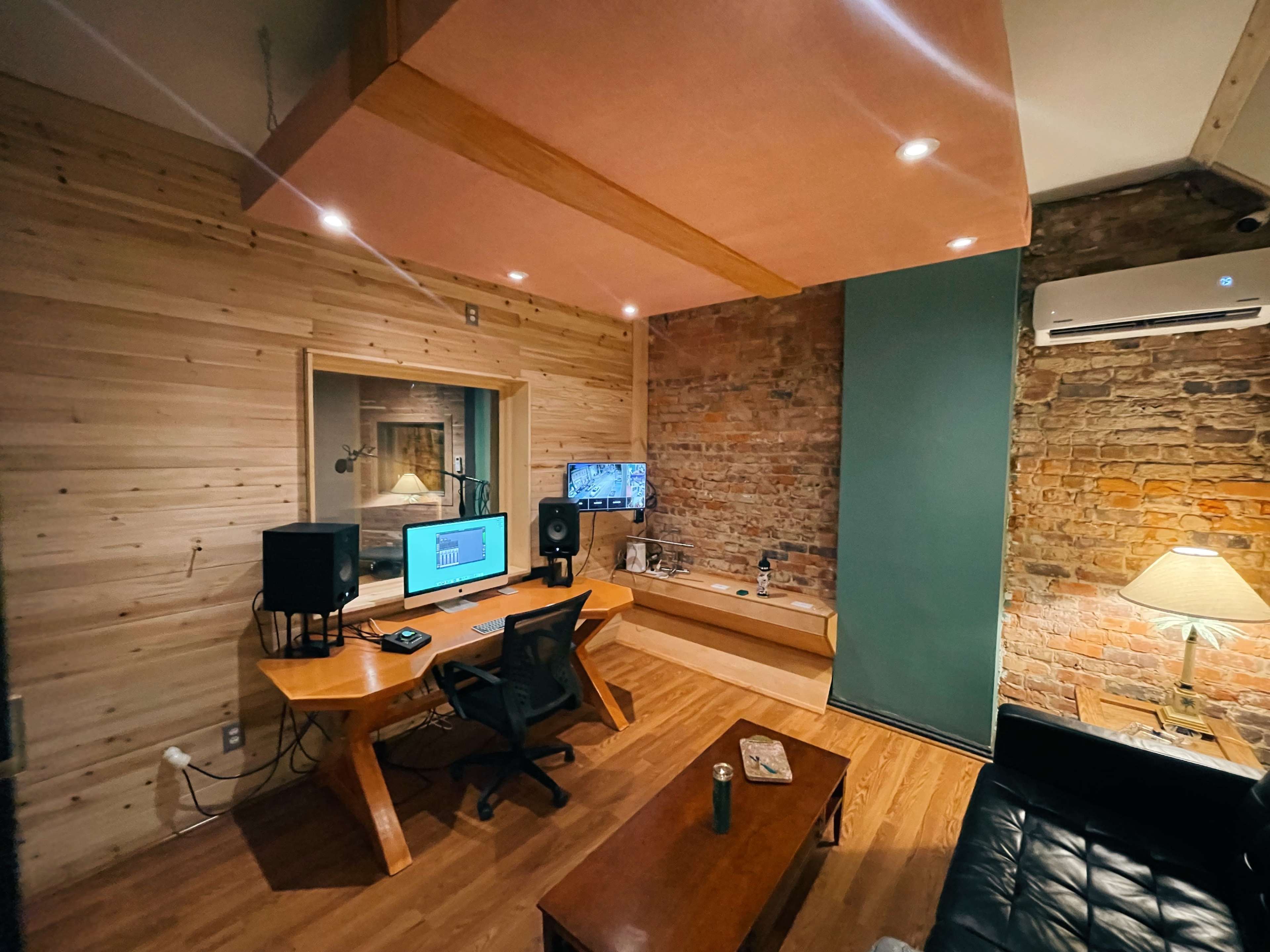 A music studio with a wooden desk, computer monitors, audio equipment, and a leather couch, featuring a mix of wooden and exposed brick walls.