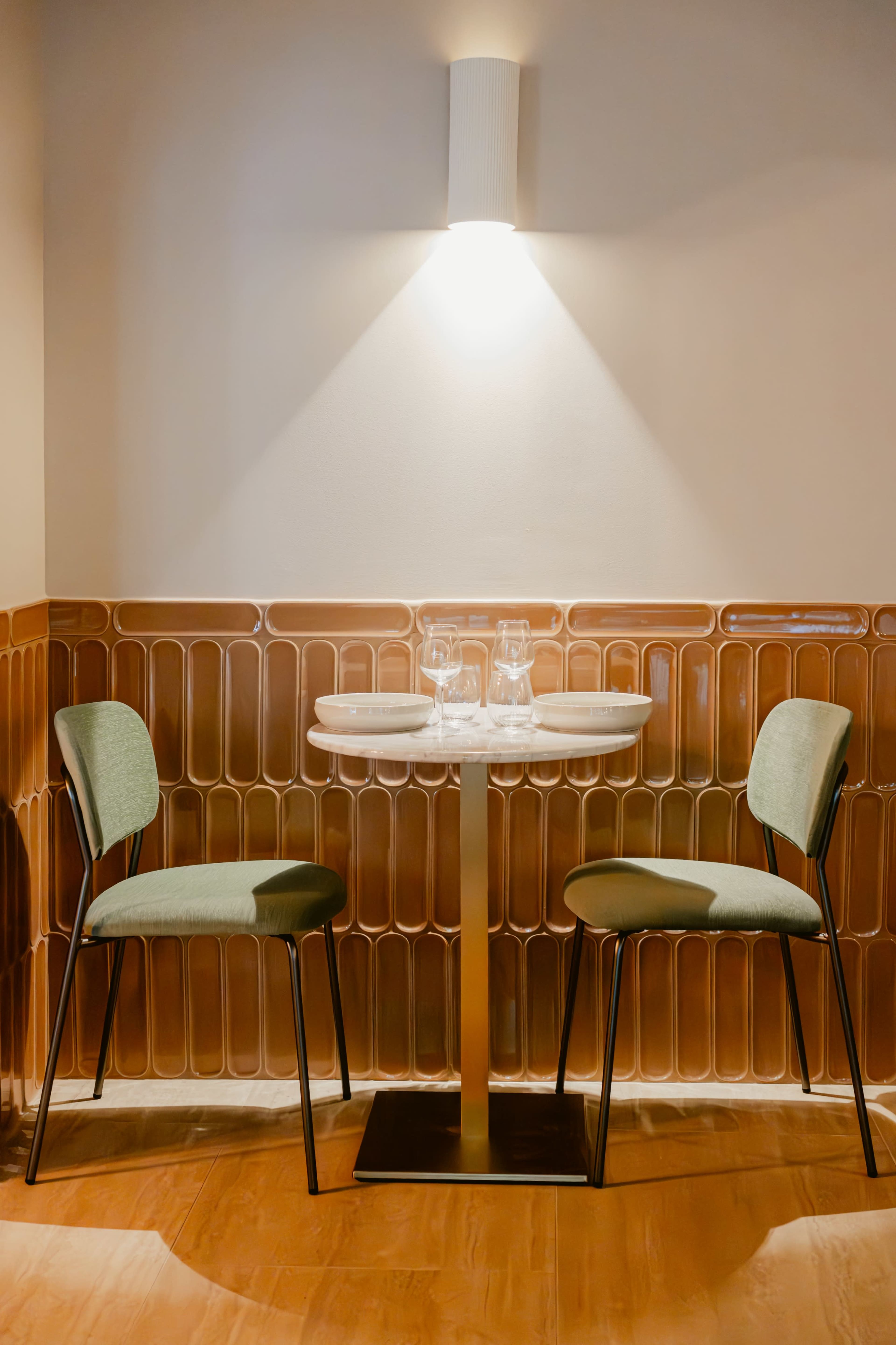 A small dining table with two chairs is positioned against a tiled wall, illuminated by a wall-mounted light.