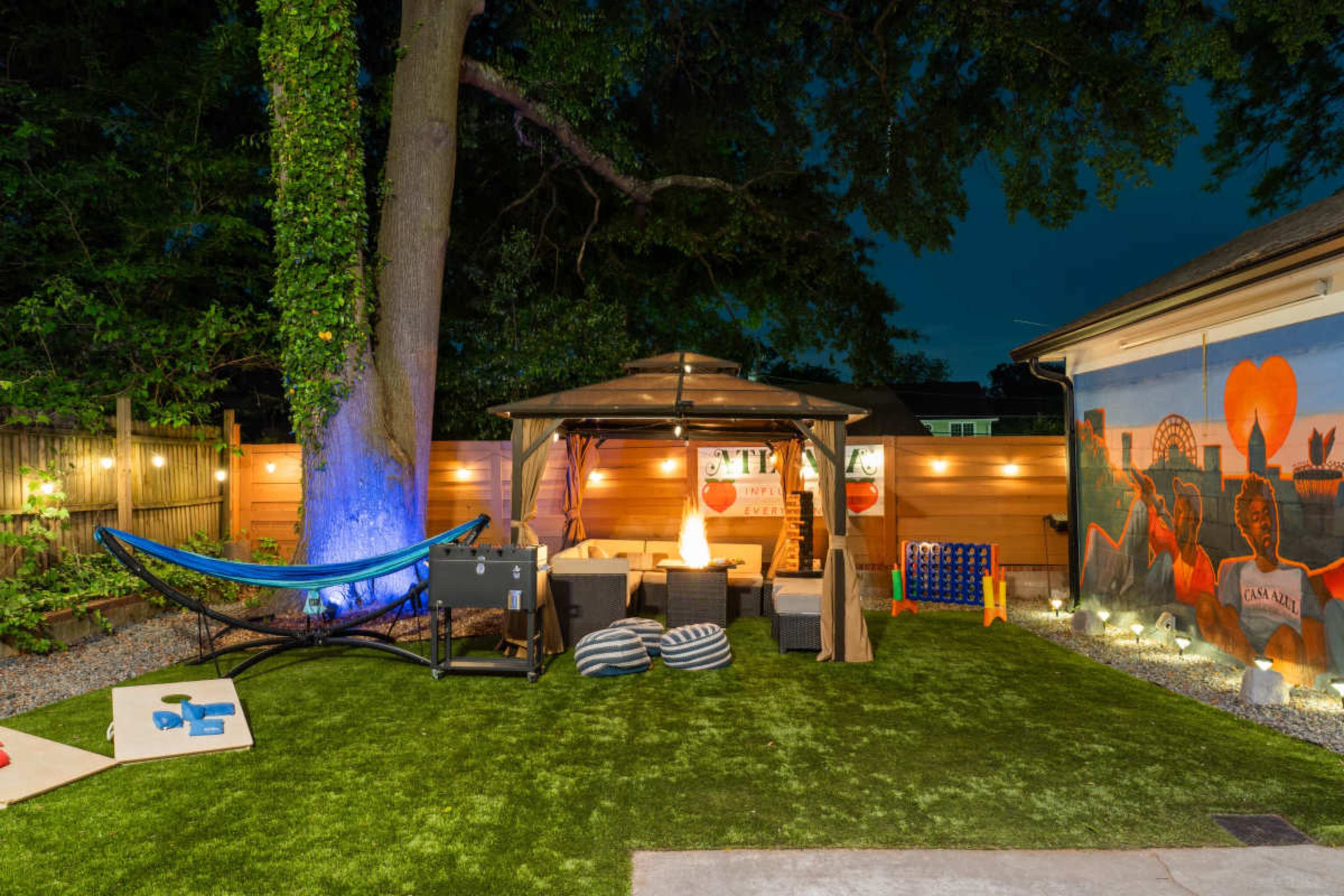 The image shows a backyard scene featuring a cozy seating area under a gazebo, a hammock, and illuminated outdoor decor with a mural on the adjacent wall.
