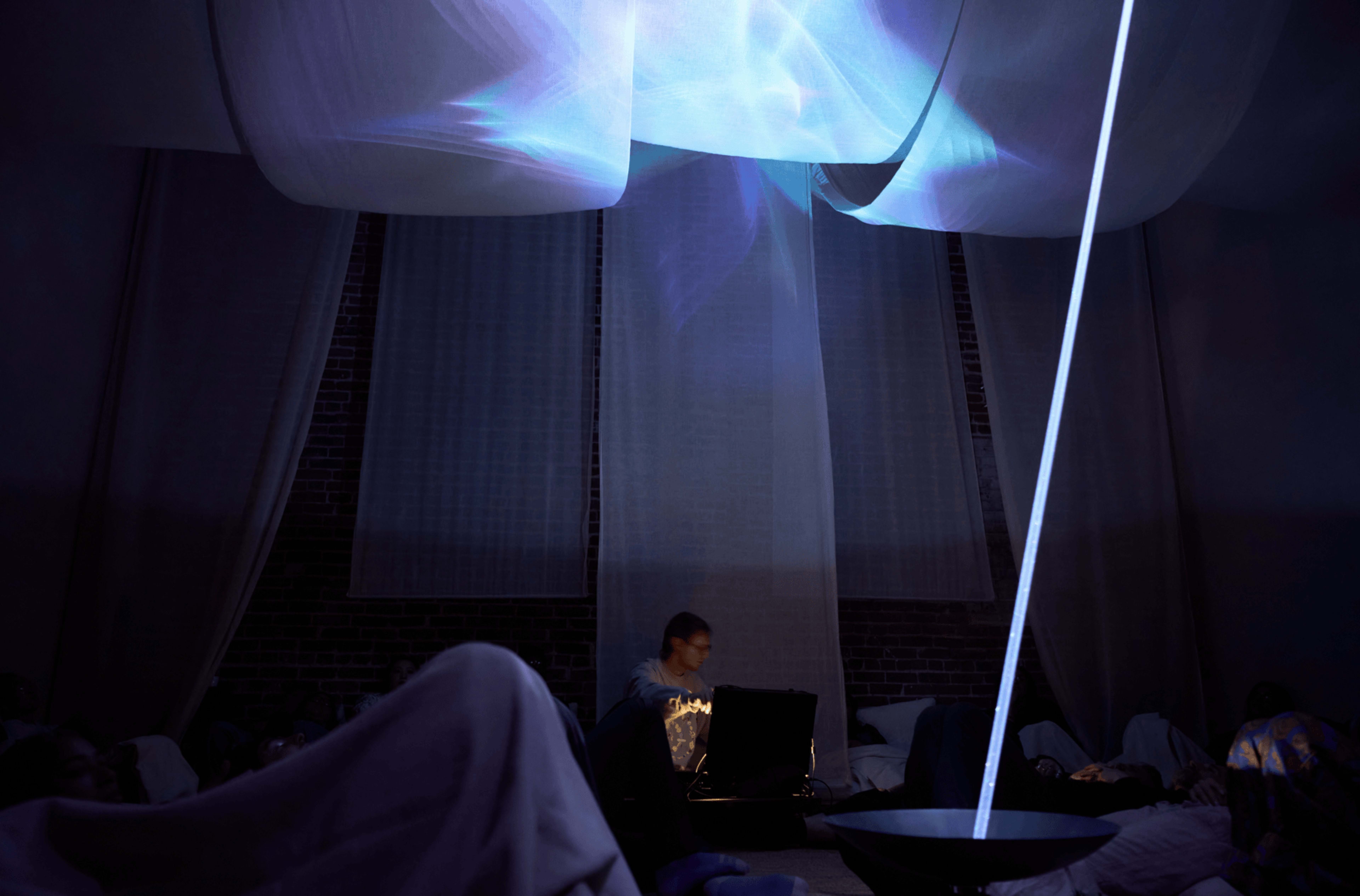 A person is sitting on the floor in a dimly lit room, operating a laptop while a projector displays colorful light patterns on a fabric canopy overhead.