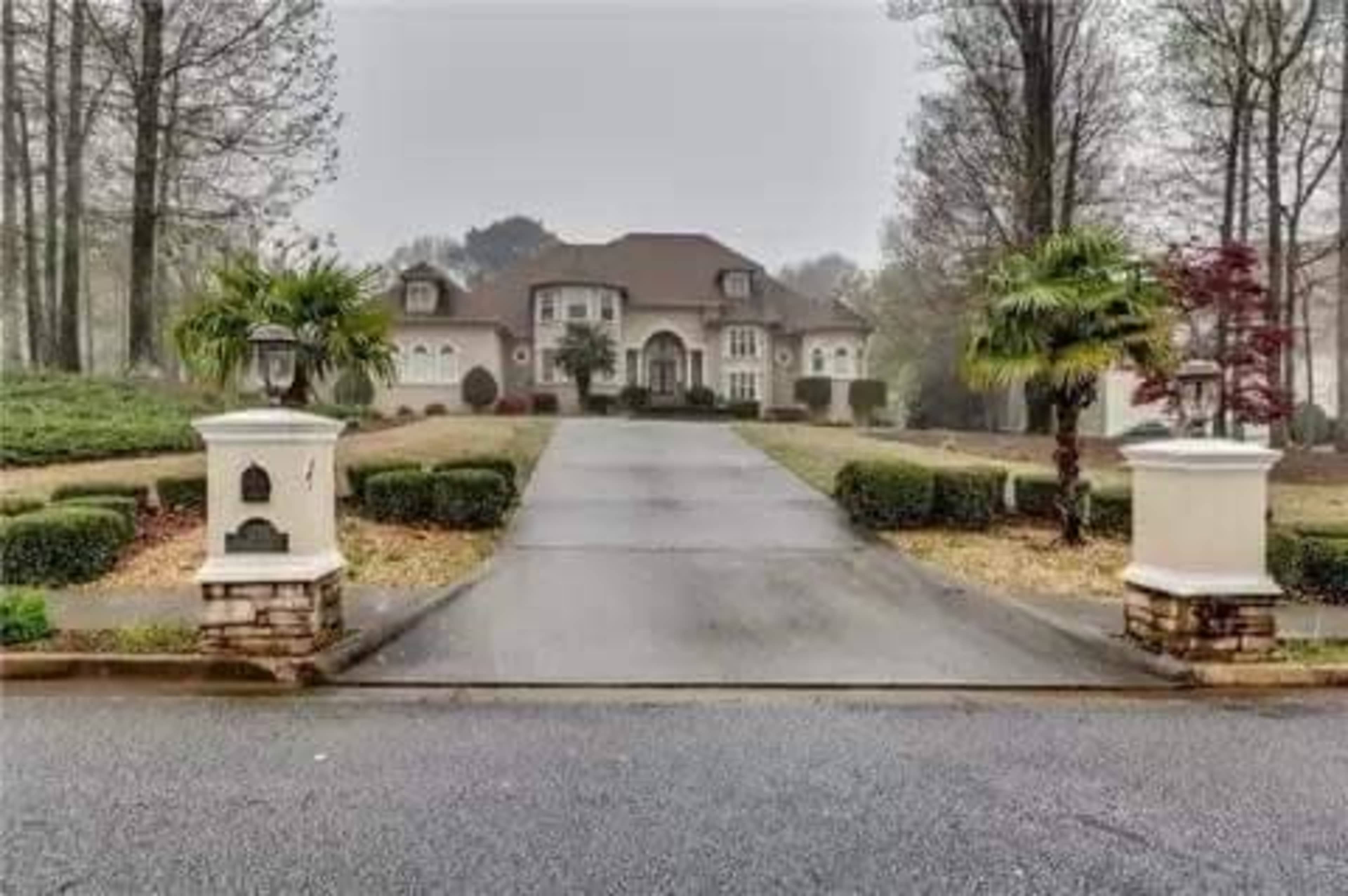 A large, upscale house set back from the road with a long driveway, flanked by palm trees and landscaped greenery.