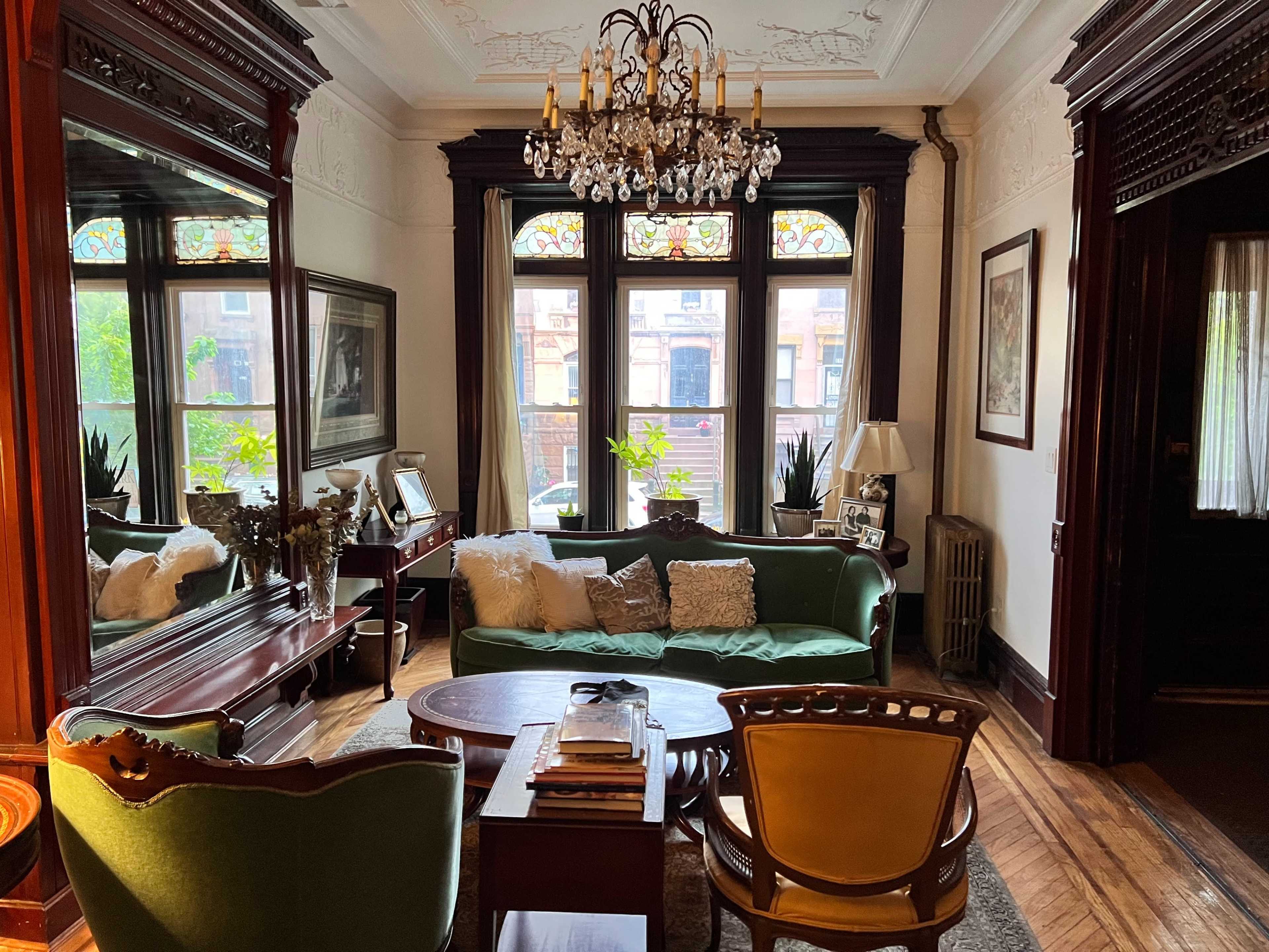 The room features a green sofa, a round coffee table, and ornate wooden furnishings, with large windows allowing natural light to illuminate the space.