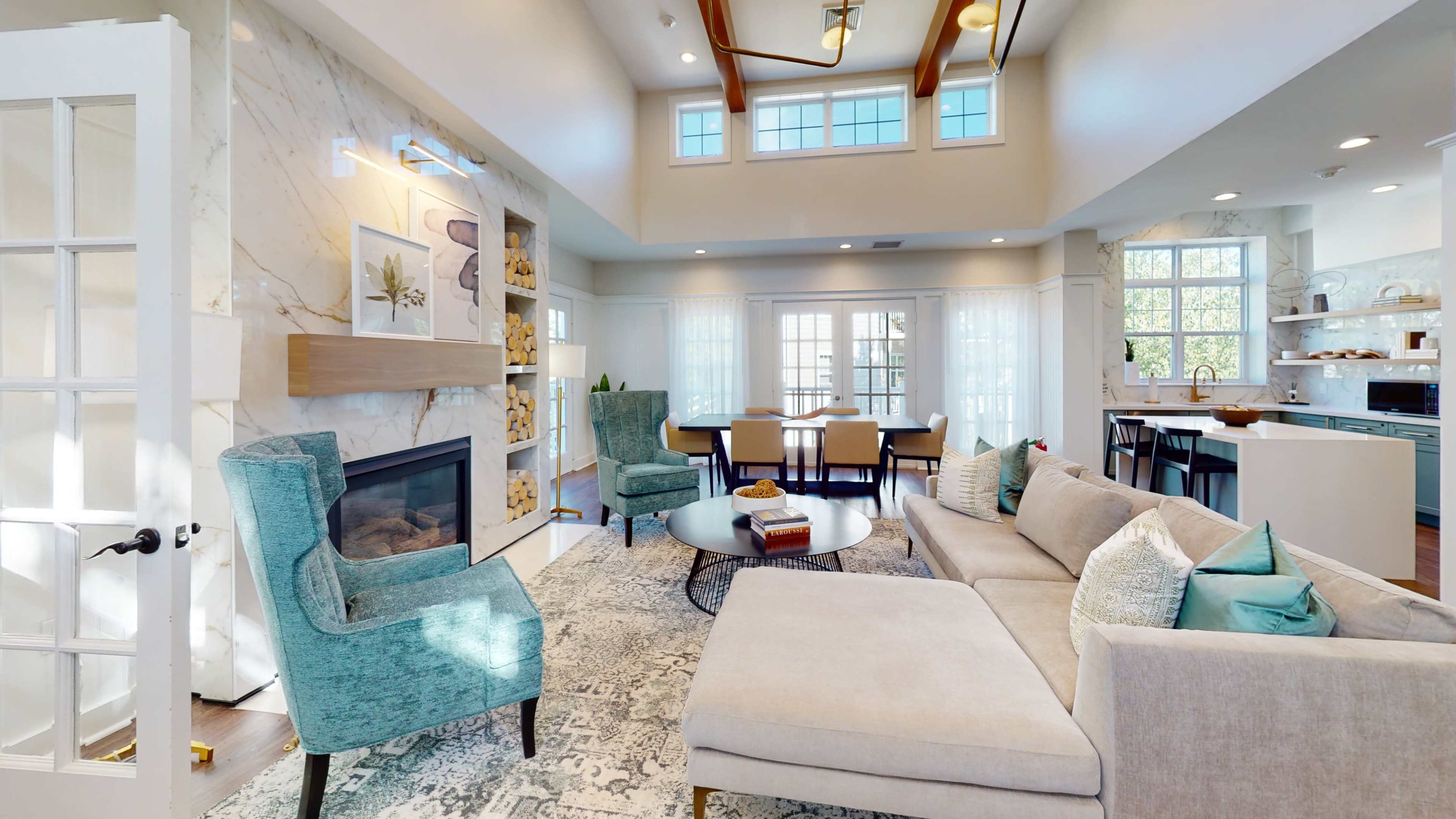 A modern living room with a sectional sofa, two blue chairs, a round coffee table, and a fireplace, all under a high ceiling with large windows allowing natural light.