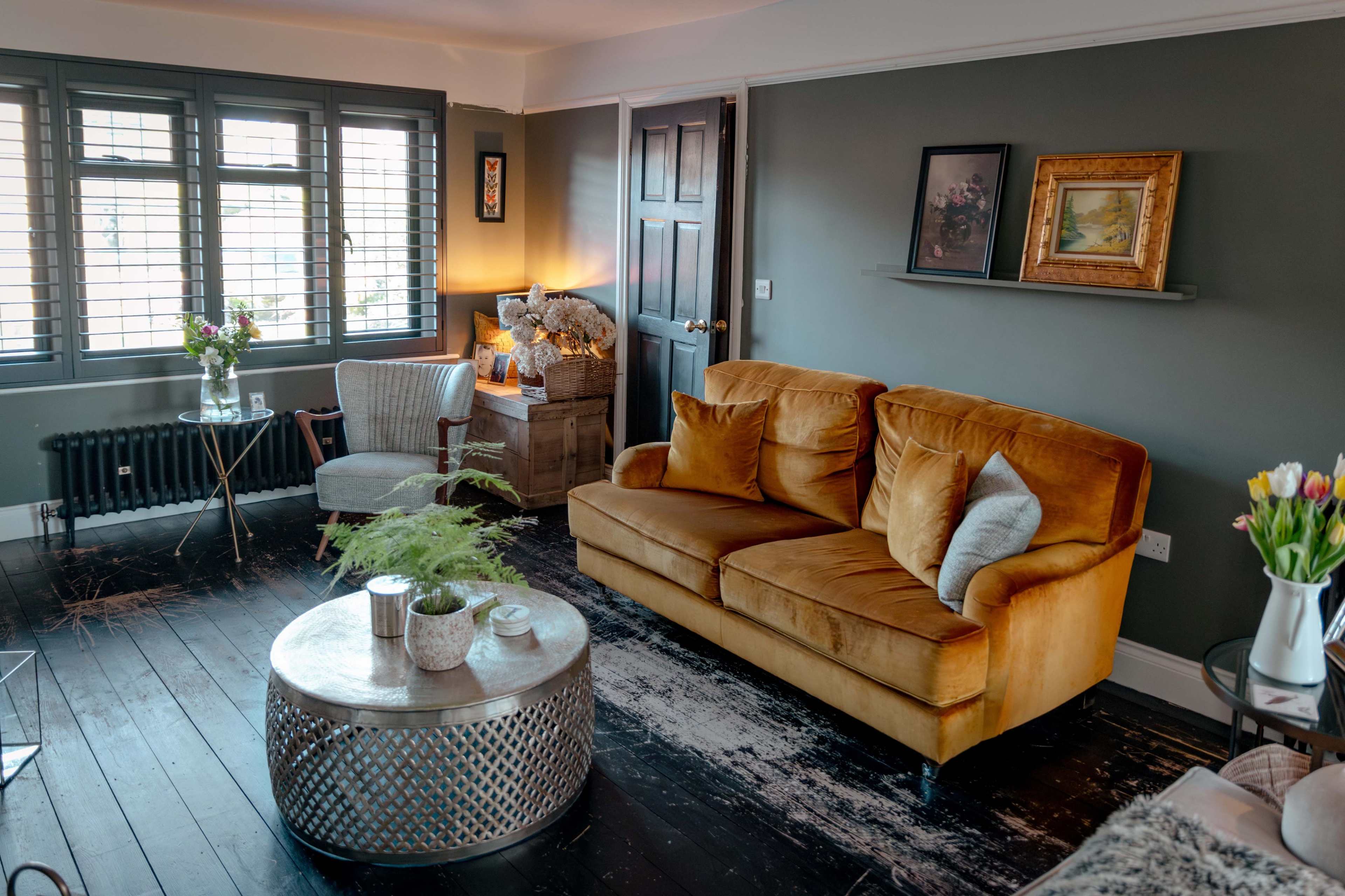The image shows a cozy living room featuring a velvet sofa, a round metallic coffee table, a chair, and a variety of decorative elements against a dark wall.