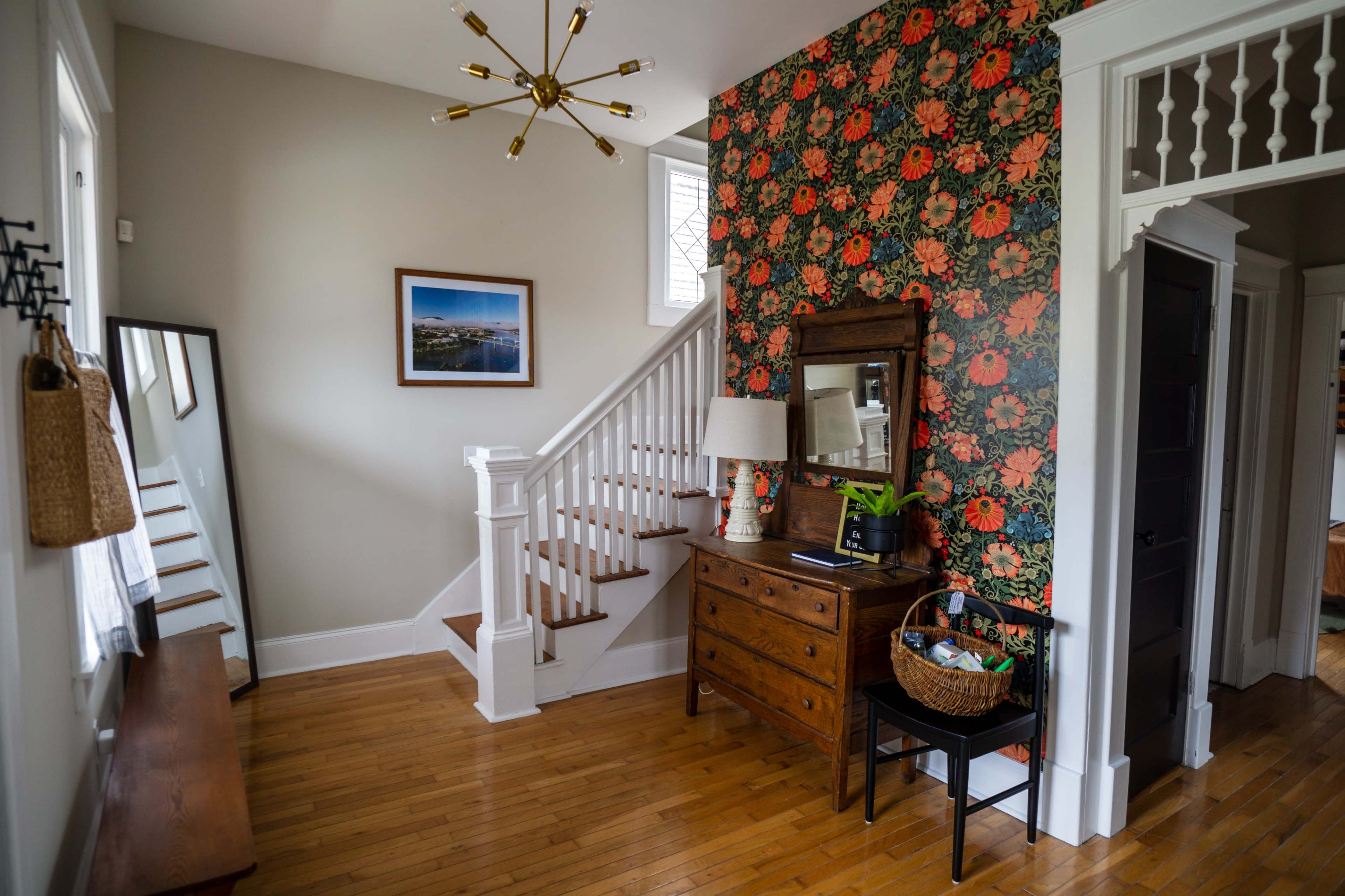 The image shows a well-lit entryway featuring a staircase, a mirror, a wooden dresser with a lamp, a floral wallpaper accent wall, and a basket on the floor.