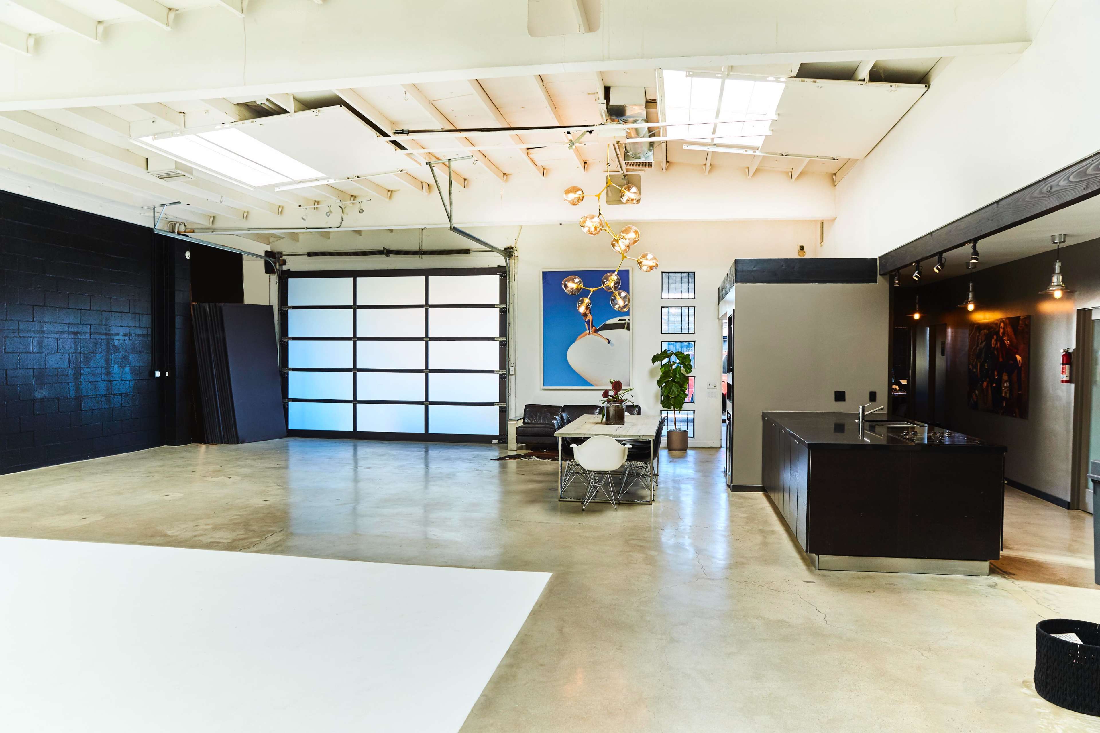 The image shows a spacious, modern interior with a large garage door, minimalist furniture, and polished concrete floors.