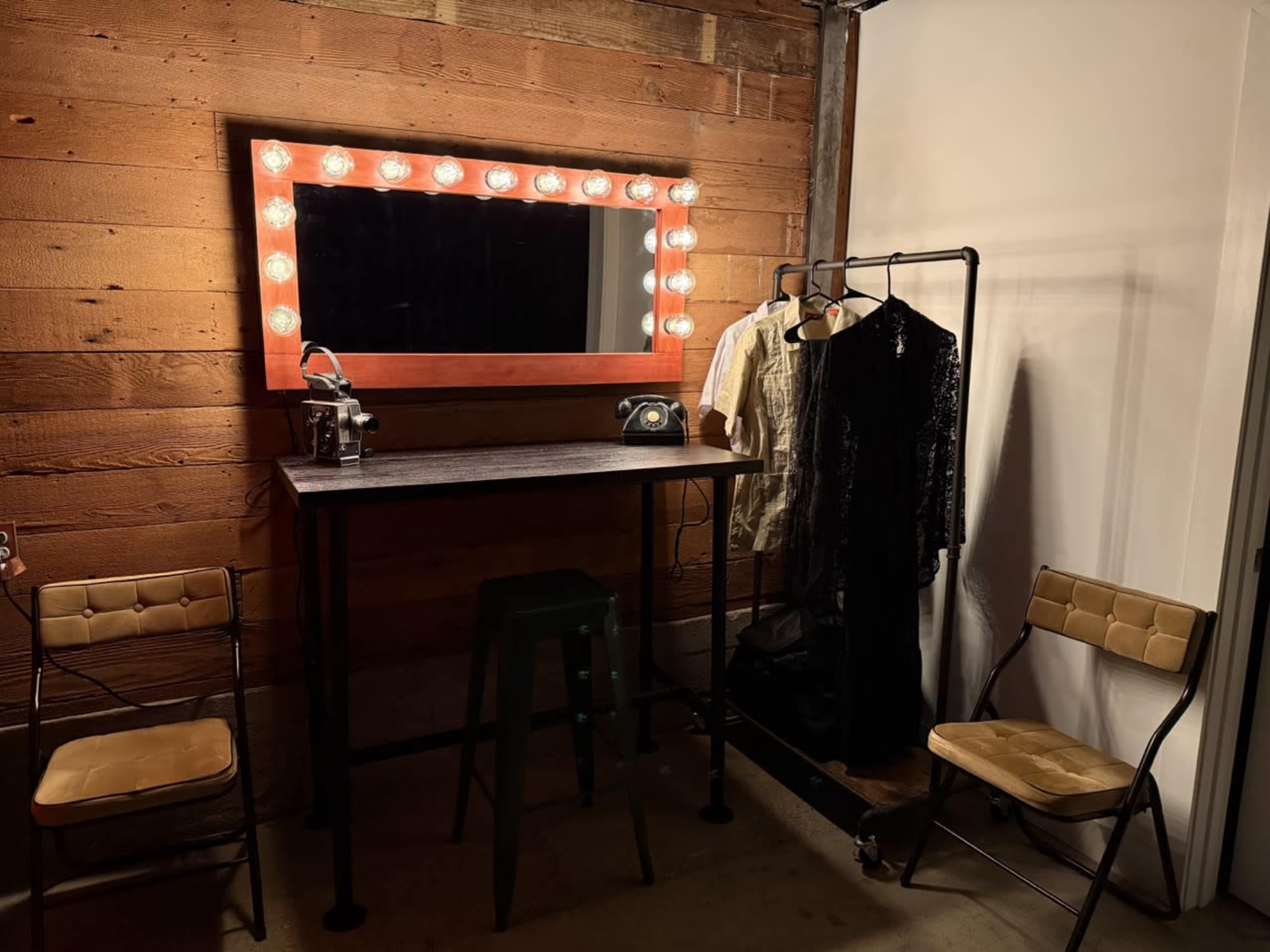The image shows a small dressing area with a mirror surrounded by lights, a vintage telephone, a clothing rack with garments, and two chairs.