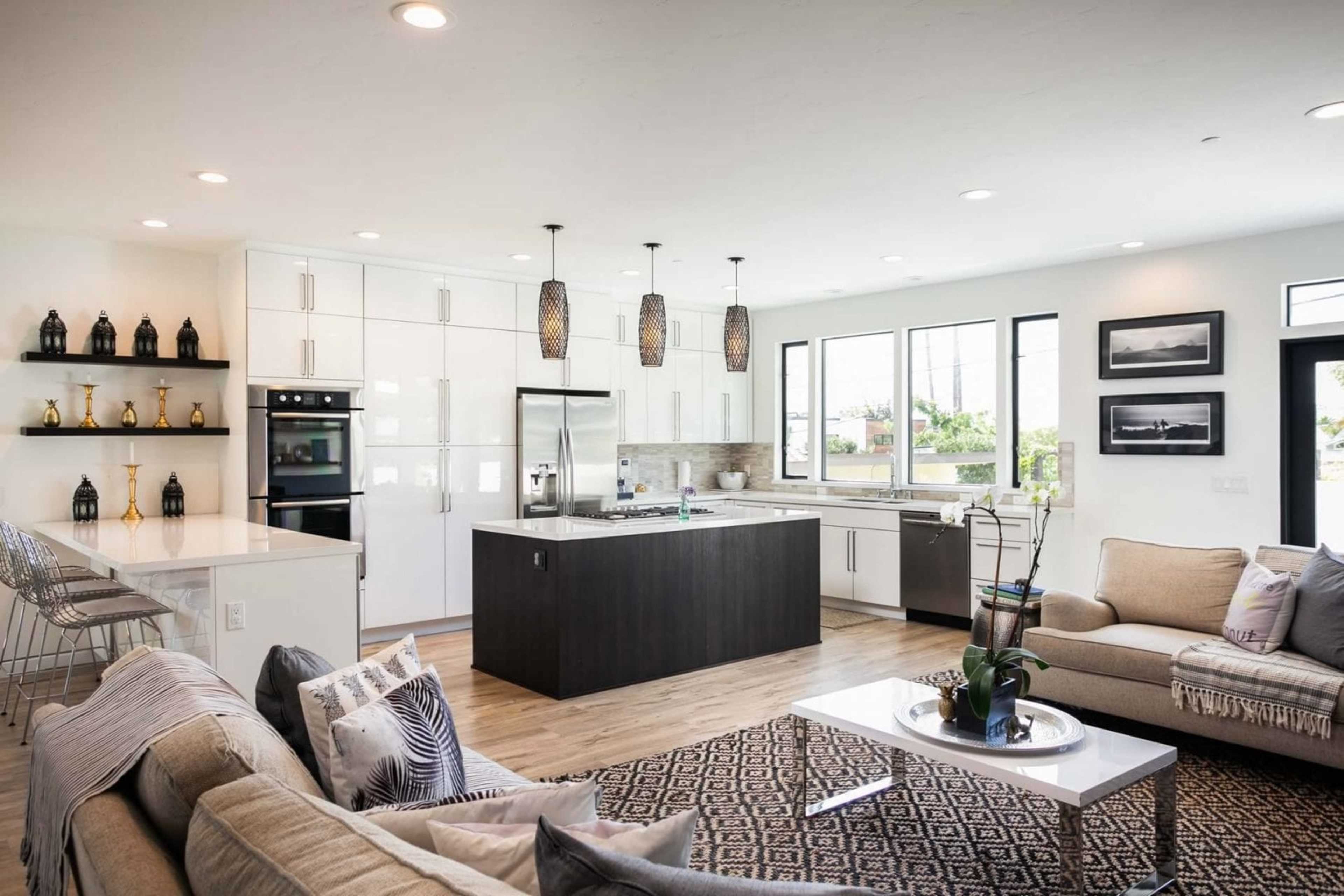 A modern kitchen and living area with white cabinetry, a central island, and large windows allowing natural light to fill the space.