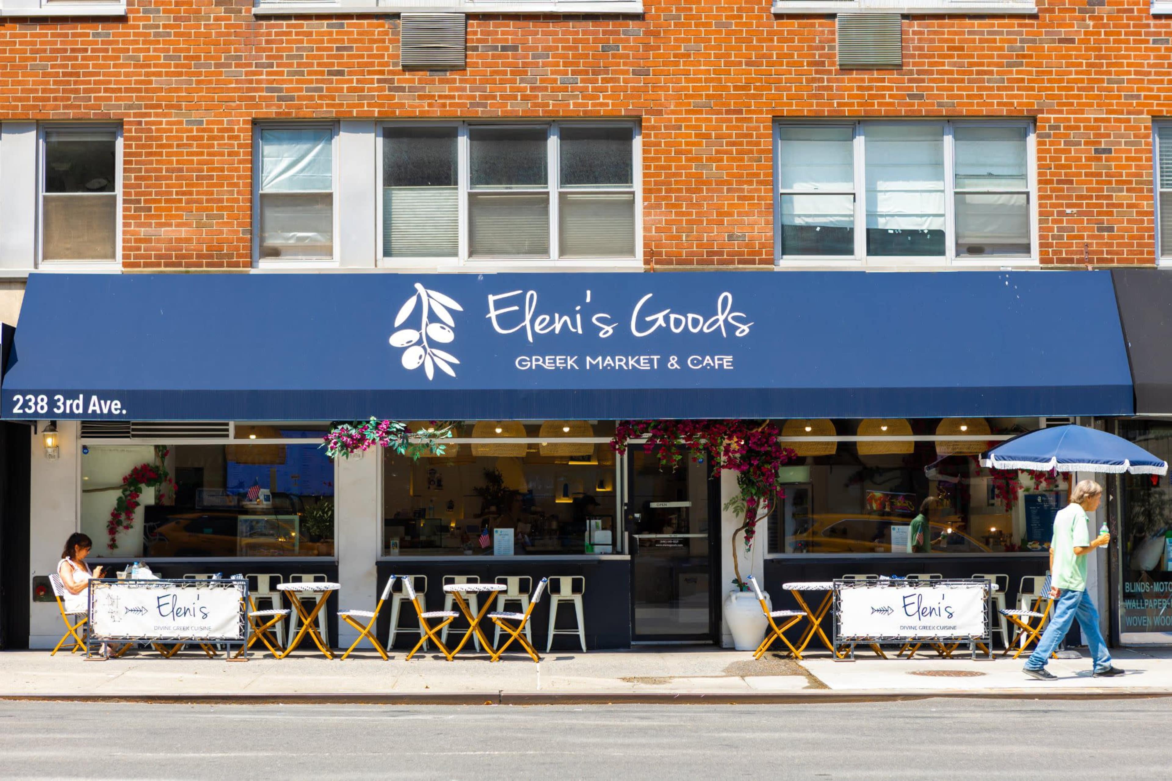 The image shows the storefront of "Eleni's Goods," a Greek market and café, featuring outdoor seating and colorful decorations.