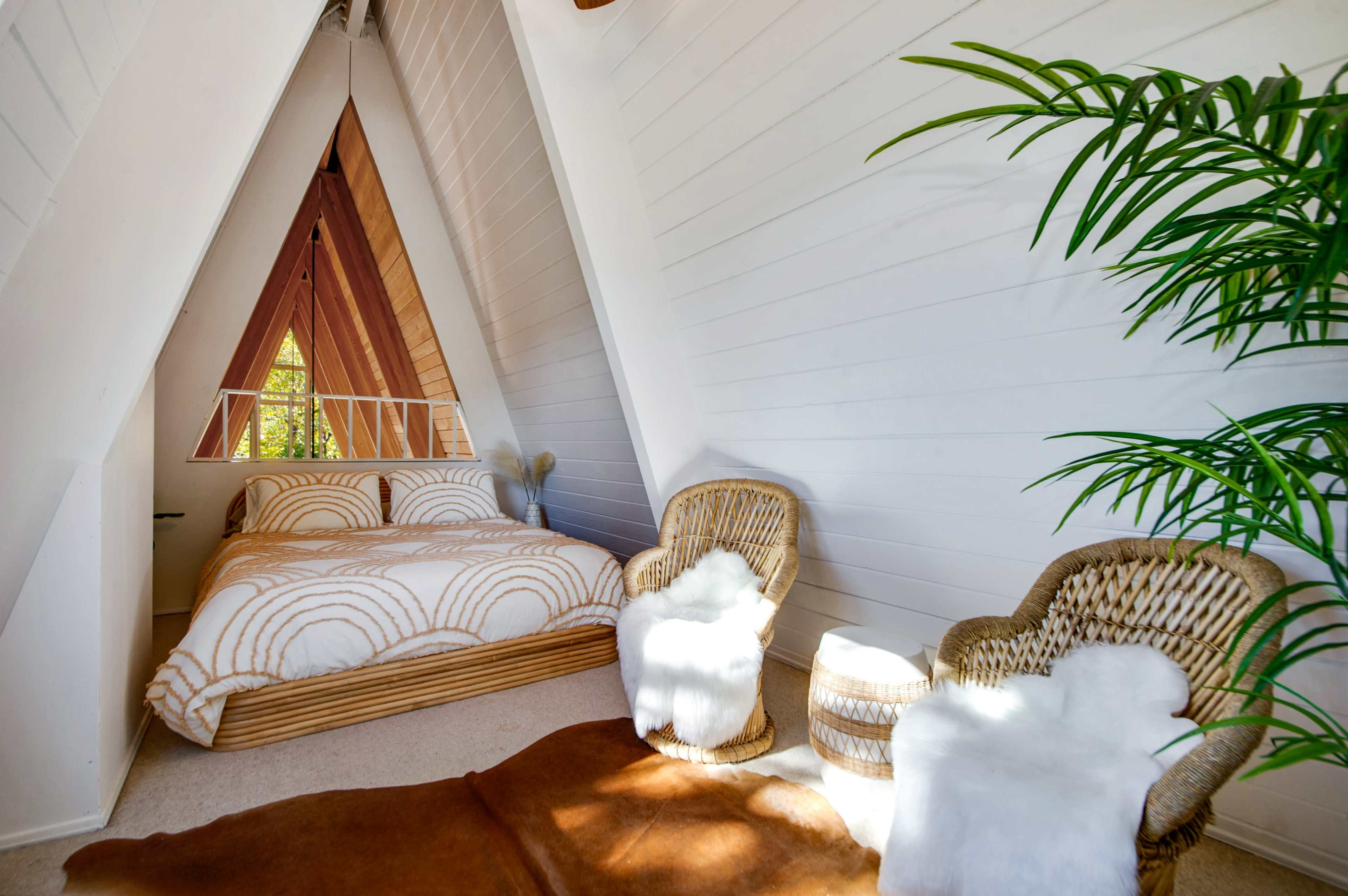 A minimalist A-frame bedroom features a central bed with patterned bedding, flanked by two woven chairs and a small table, set against white walls and wooden accents.
