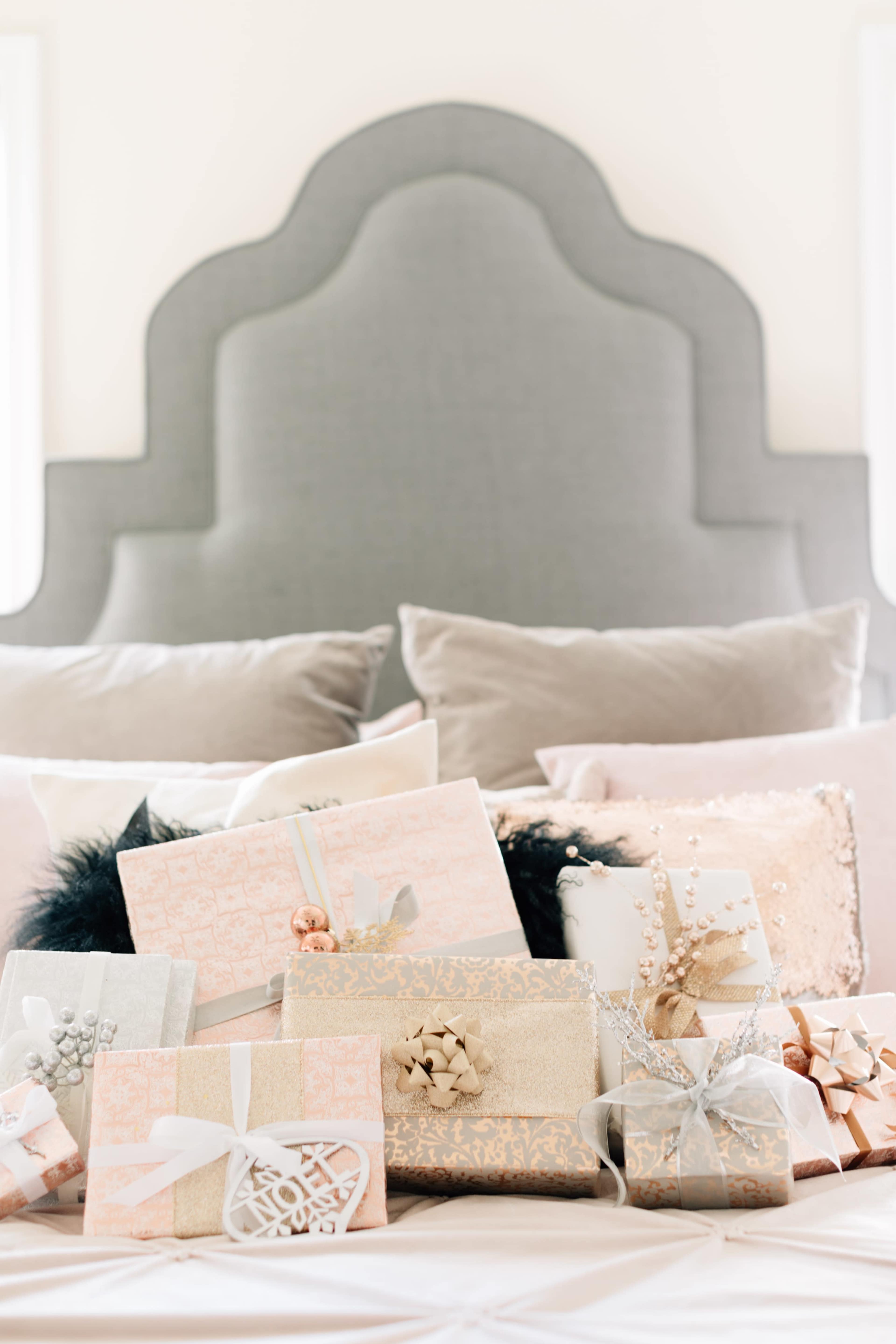 The image shows a neatly arranged collection of wrapped gifts on a bed with a large, upholstered headboard and decorative pillows in the background.