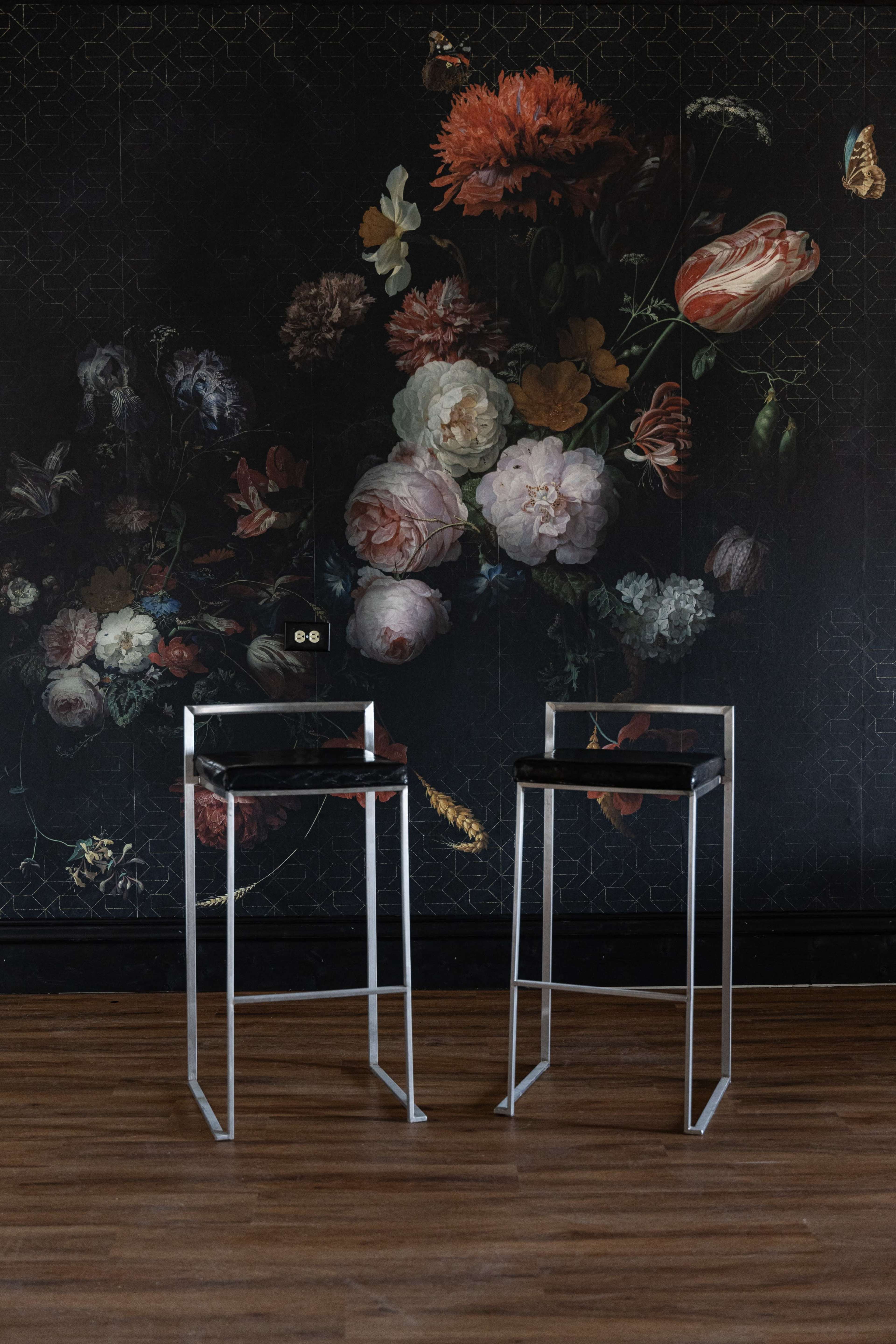 Two modern metal bar stools are positioned in front of a dark floral mural featuring various flowers on a wall with a wooden floor.