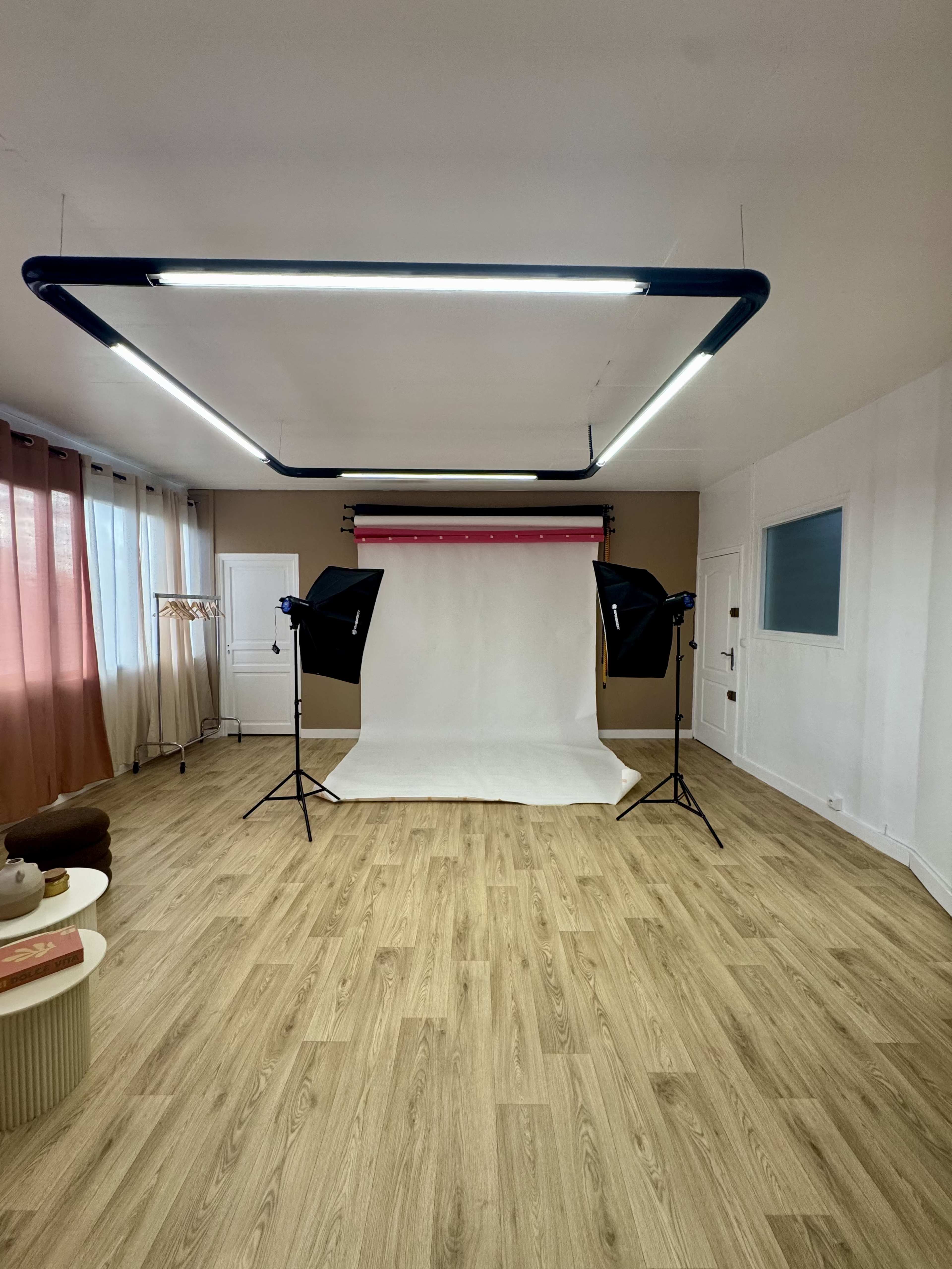 The image shows a photography studio with a white backdrop, two softbox lights, and a wooden floor, surrounded by curtains and minimal furniture.