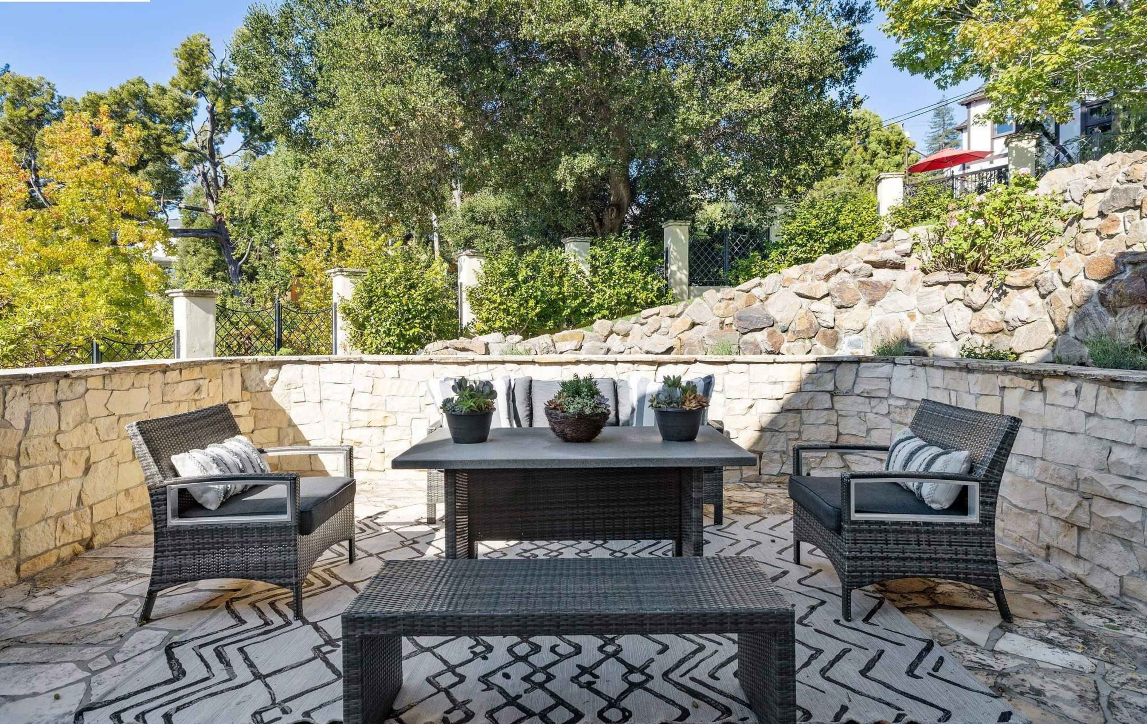 The image shows a stone patio area with a rectangular table and four wicker chairs, surrounded by greenery and a stone wall.