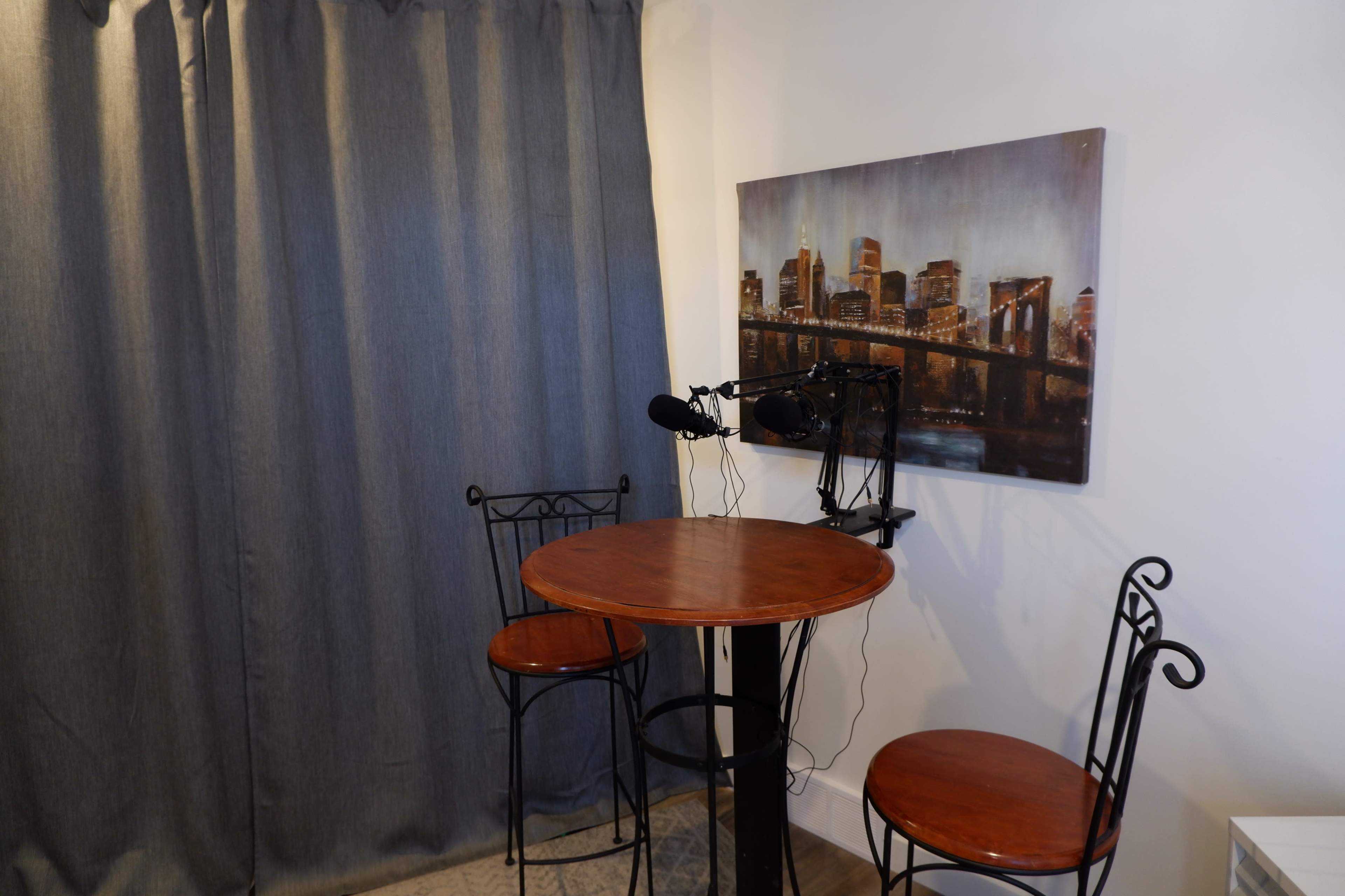 The image shows a small room with a round wooden table and two metal chairs, alongside a wall-mounted microphone and a framed cityscape painting.