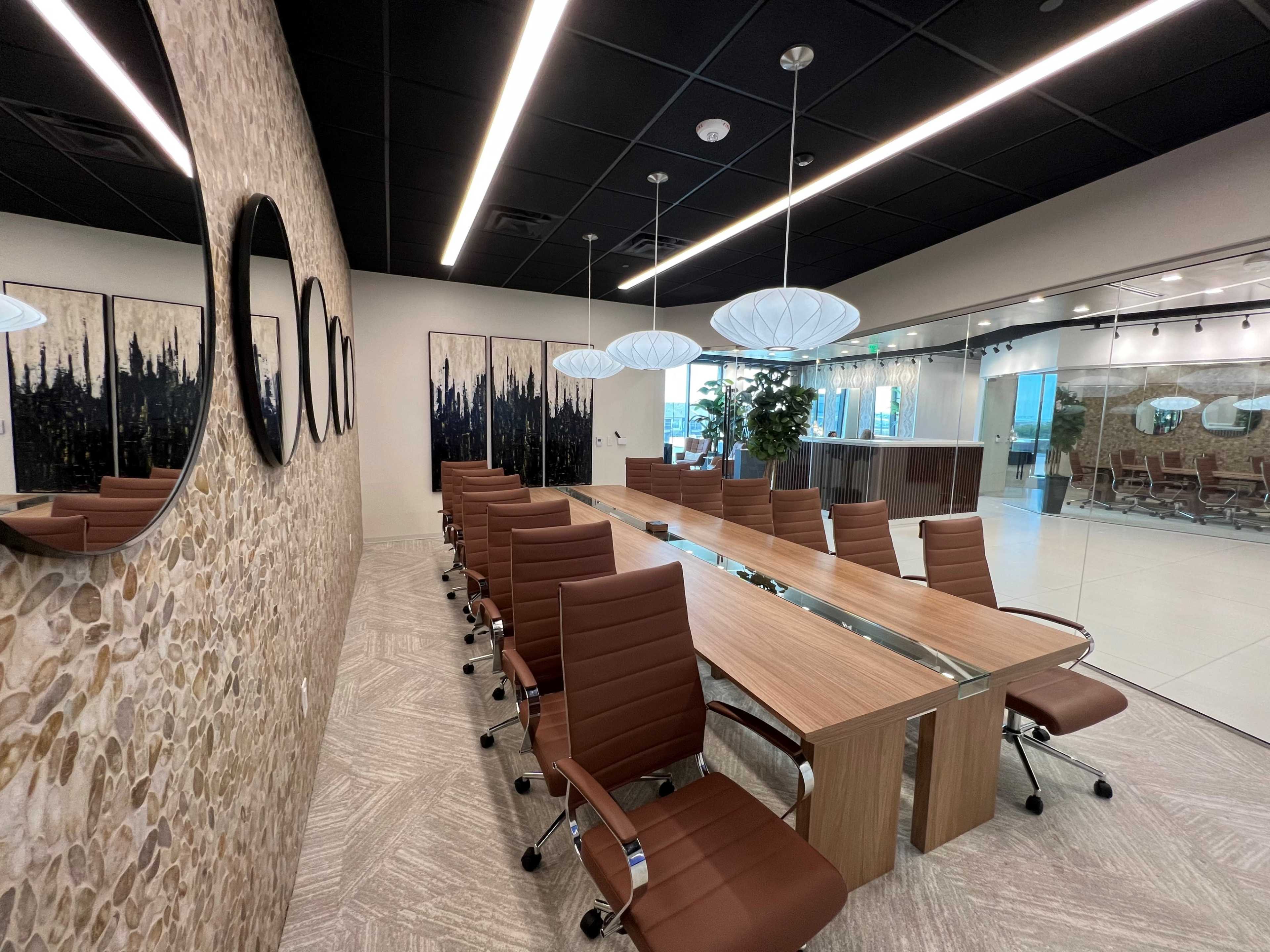 A modern conference room features a long wooden table surrounded by brown leather chairs, with stone-textured walls and large mirrors, complemented by artistic wall panels and overhead lighting.